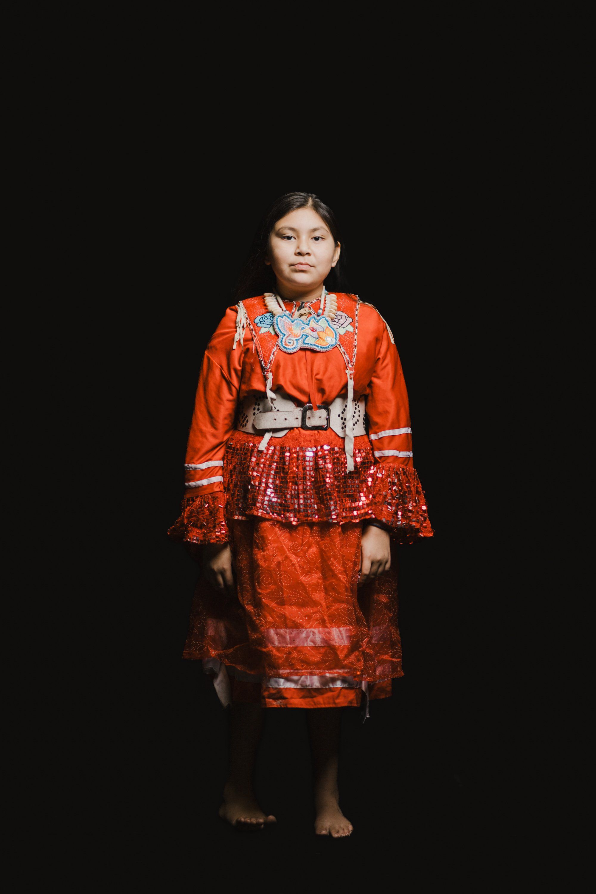 Young person in vivid orange Coast Salish regalia with geometric patterns stands against black background, barefoot and poised