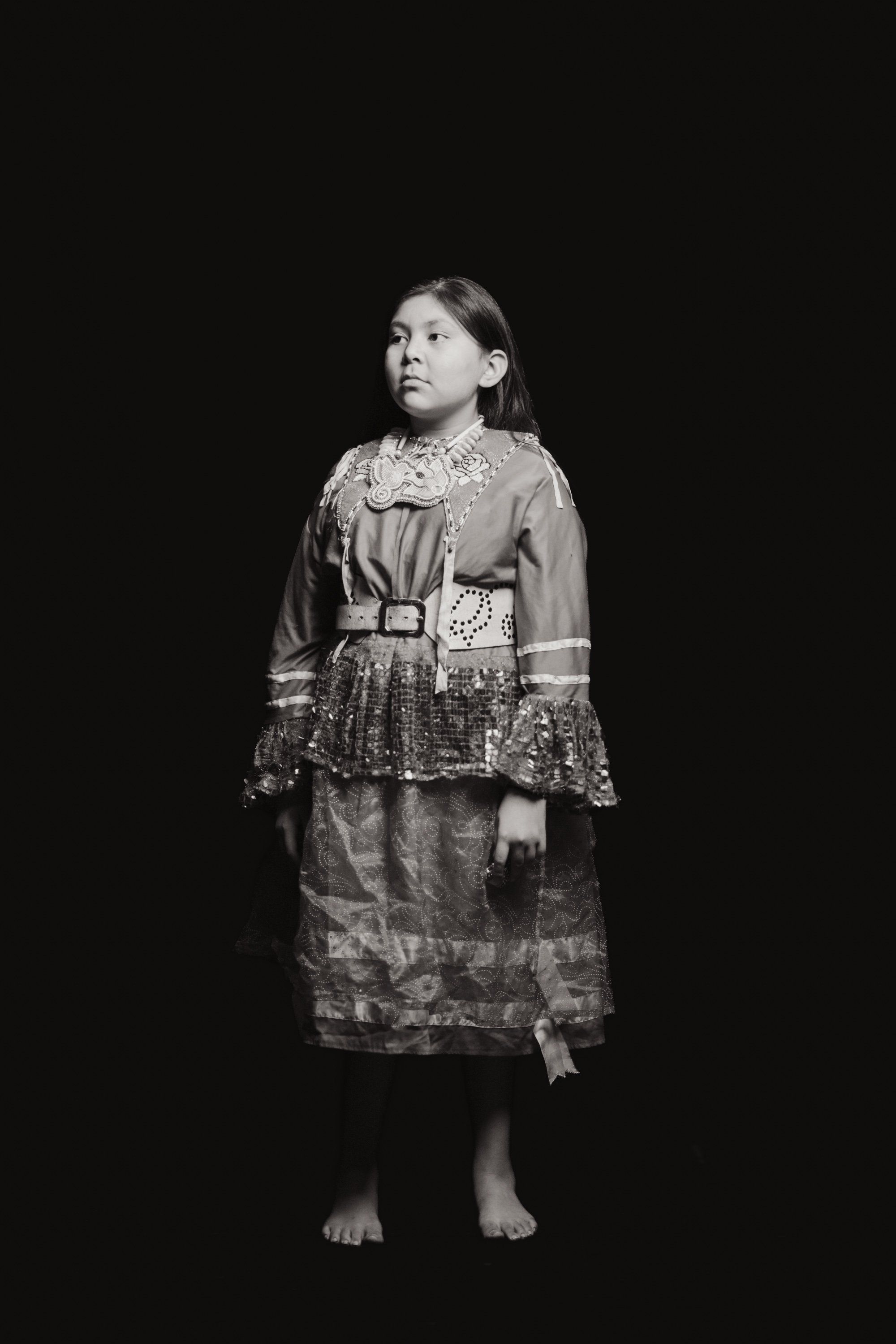 Young girl in traditional Coast Salish dress with decorative blanket and beaded details, gazing upward against dark background