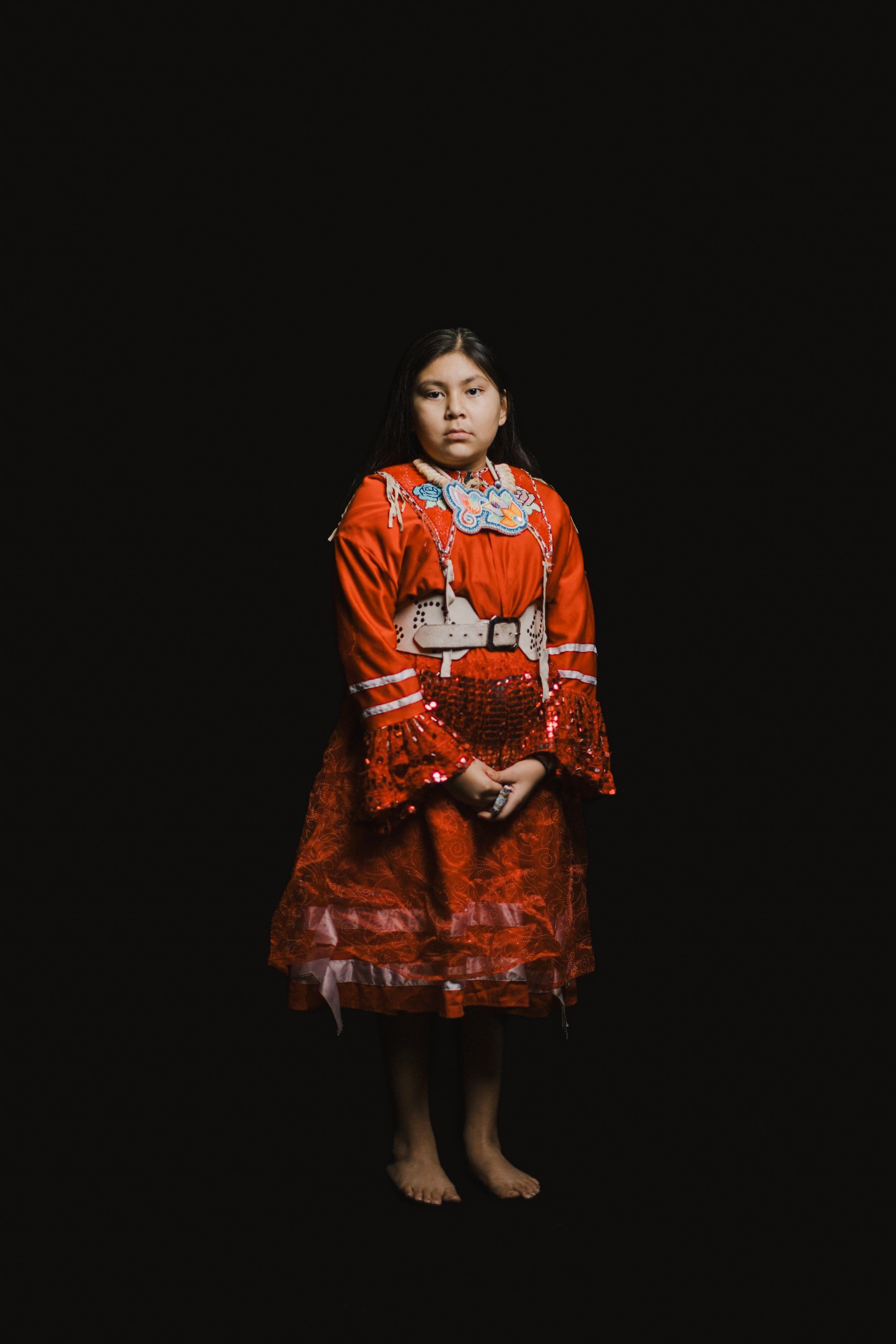 Young girl in vibrant orange Coast Salish regalia stands barefoot against black background, gazing directly at camera