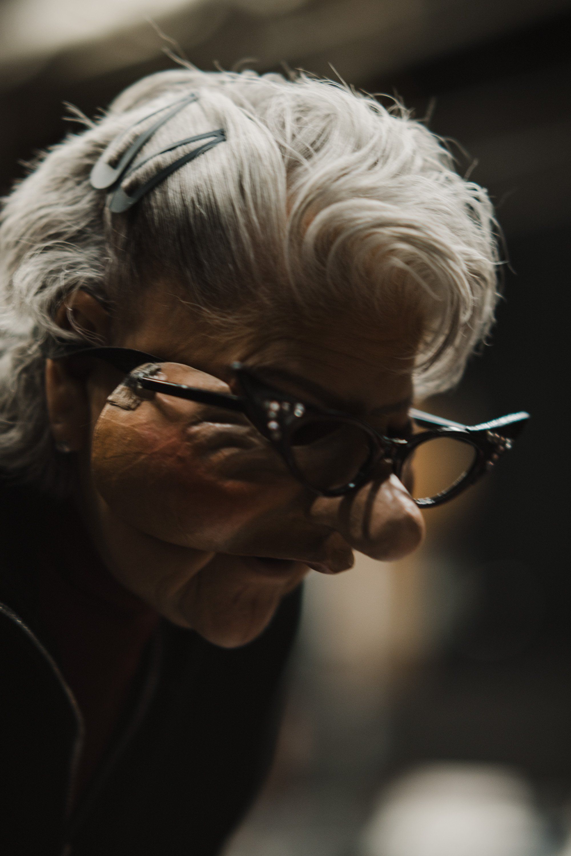 Elderly woman with silver hair and dark-framed glasses in profile, warm sidelight against dark background