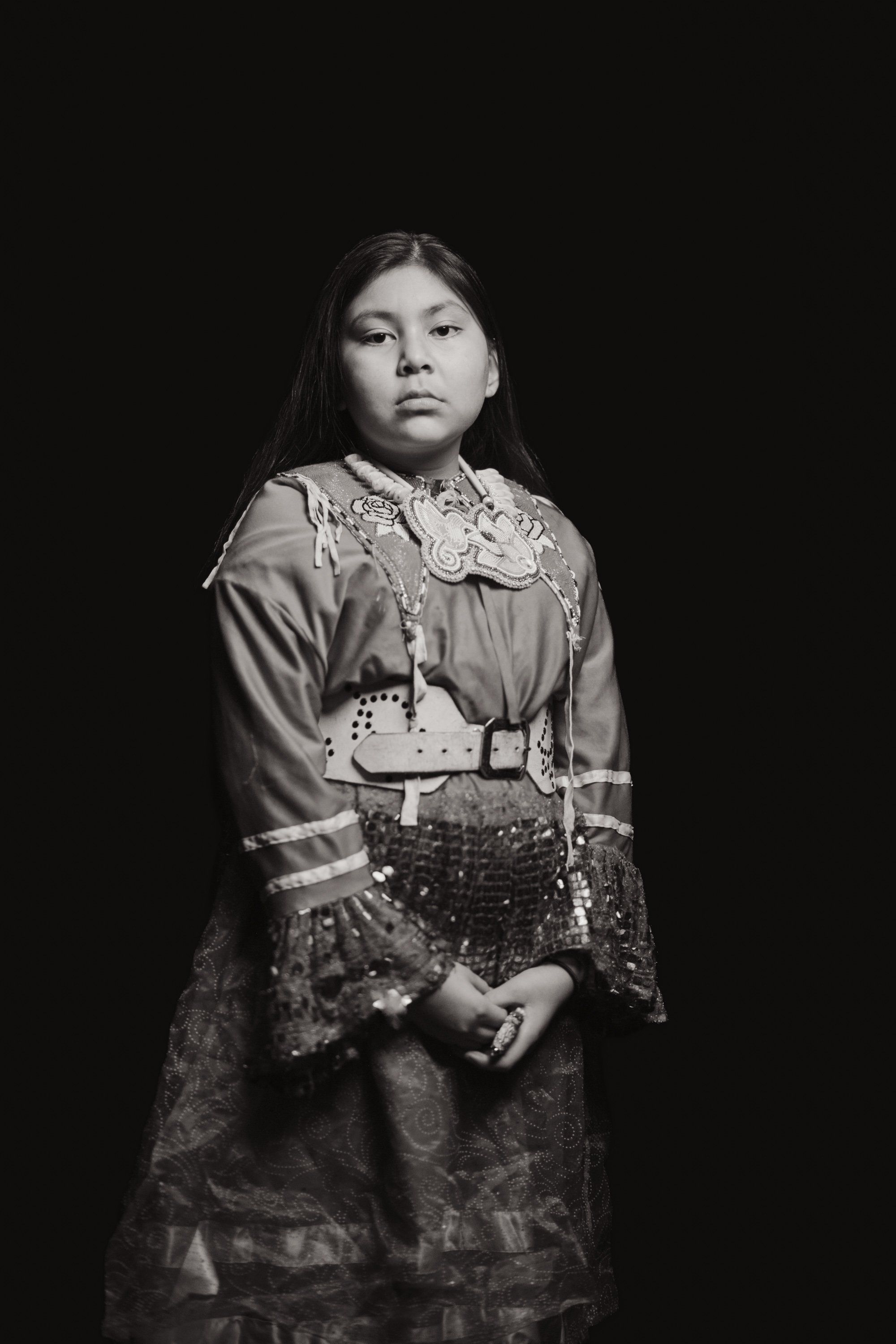 Young person in traditional Coast Salish regalia with ornate beaded belt and lace collar, posed against dark background with contemplative expression