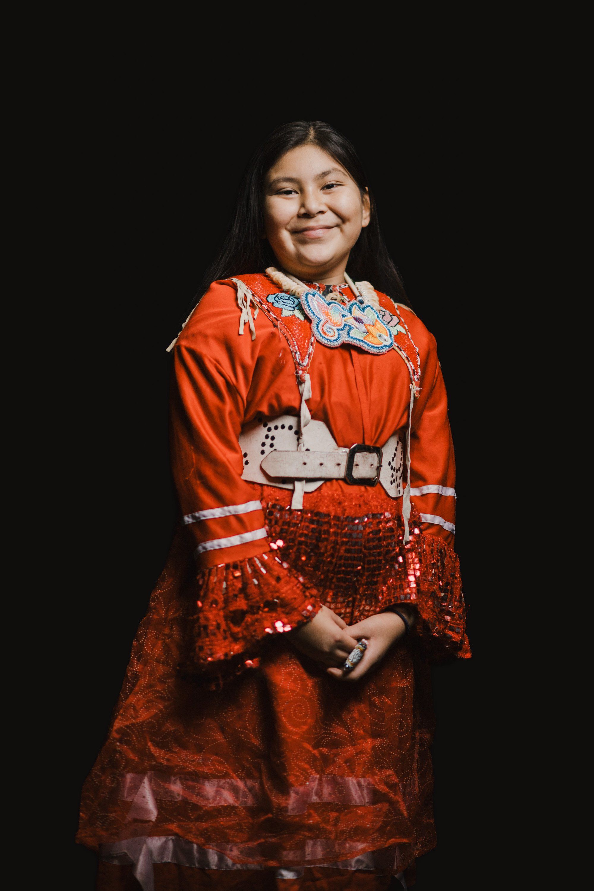 Young girl in vibrant orange Coast Salish regalia with white geometric designs smiles warmly against dark background