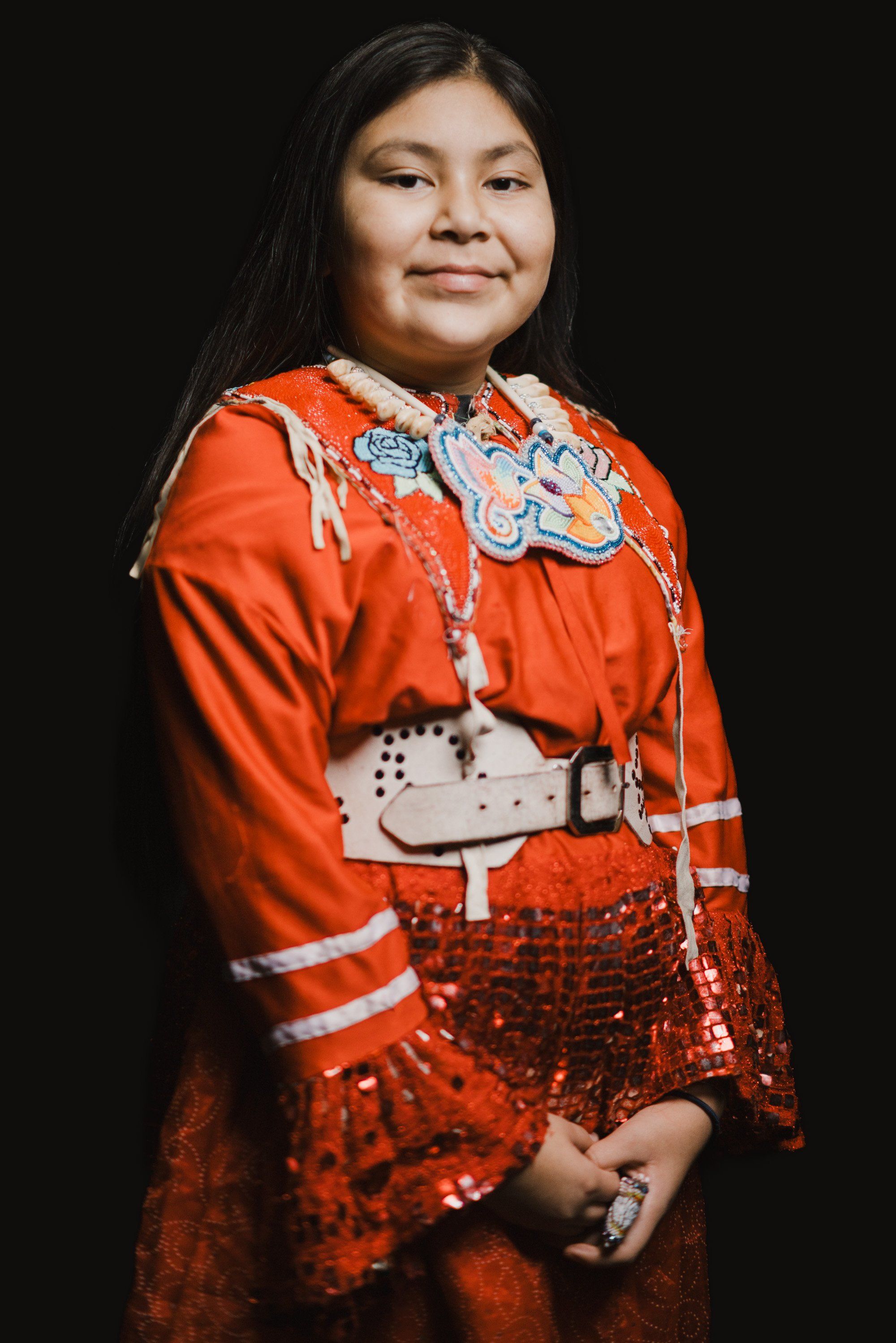 Woman in vibrant orange regalia with beaded appliqué and sequined skirt smiles warmly against black background, long dark hair framing her face