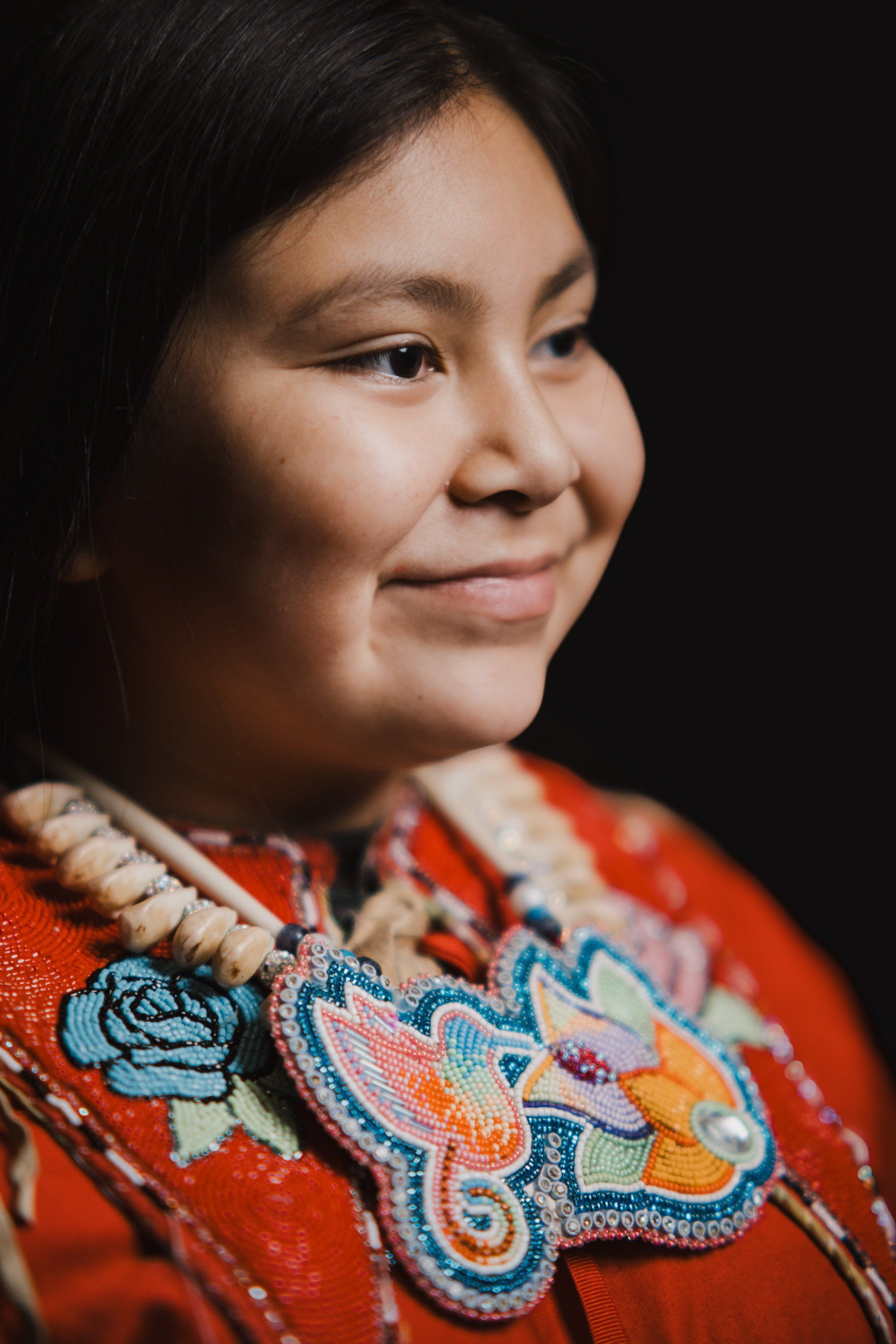 Young person in vibrant red regalia with intricate blue and gold beadwork smiles gently against a black background