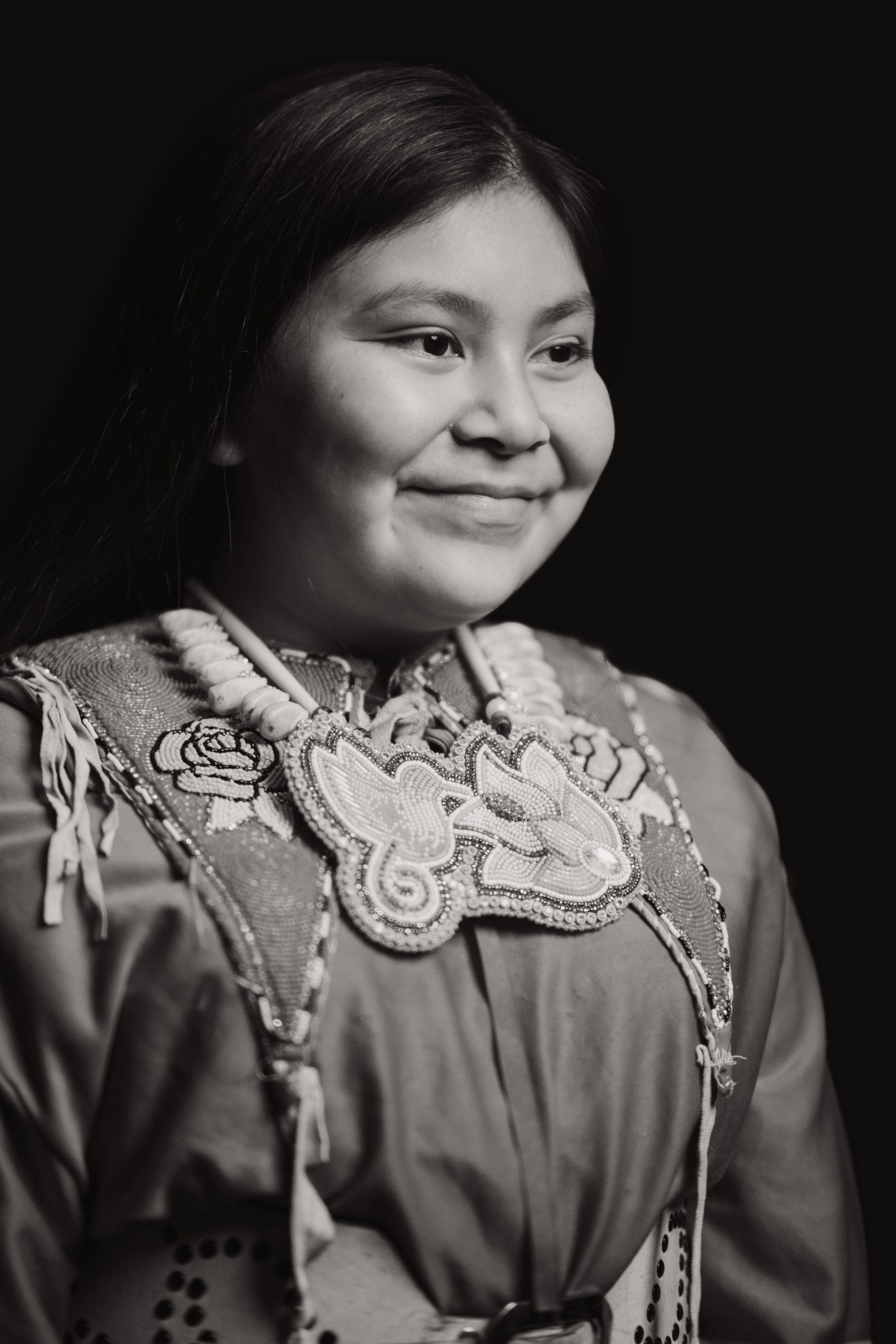Young person in traditional Coast Salish regalia with ornate beadwork, smiling warmly against dark background in studio portrait