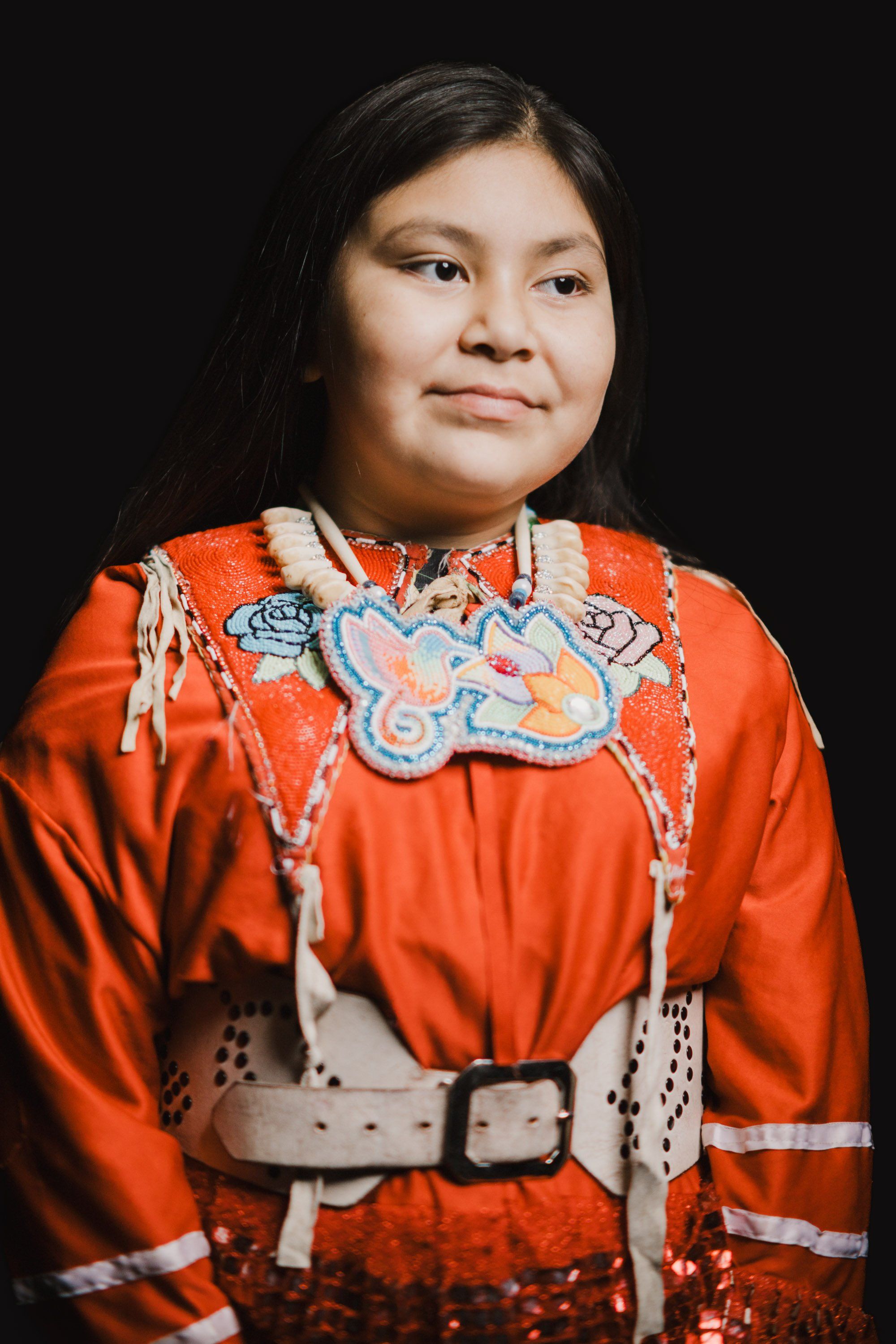 Young person wearing vibrant orange regalia with intricate blue and white beadwork, photographed against black background with warm studio lighting