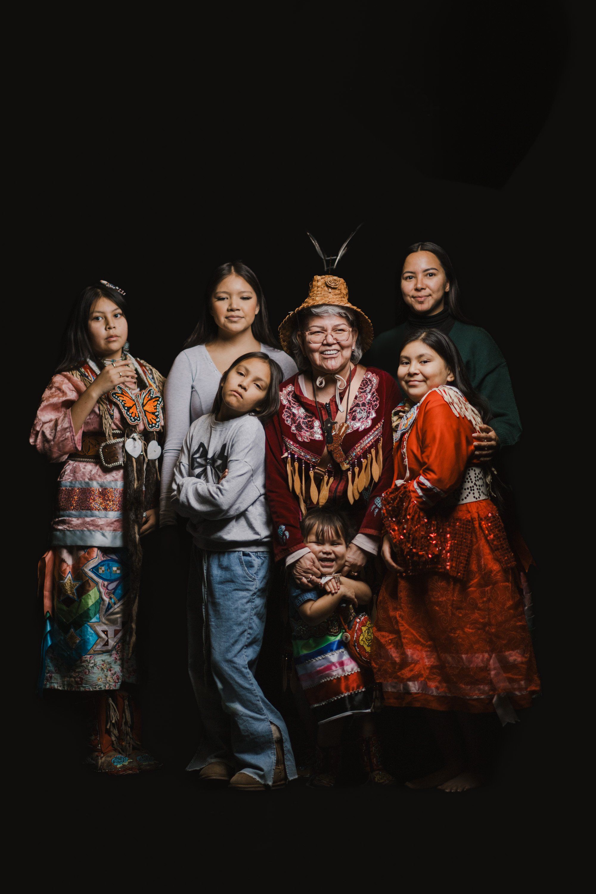 Six Coast Salish family members in traditional regalia pose against a black backdrop, their colorful blankets and clothing creating warm tones in the intimat…