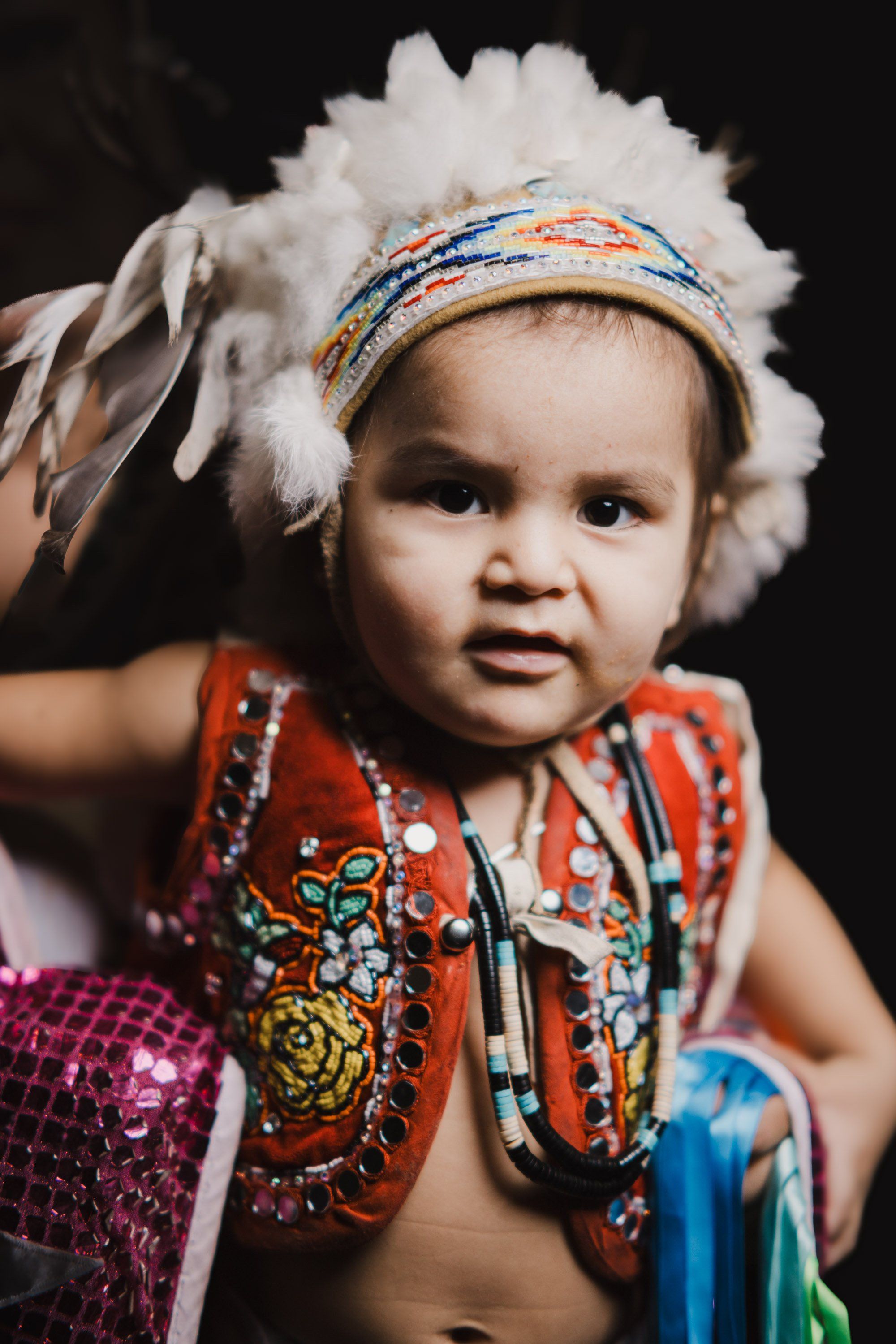 Young child in traditional regalia with white feathered headdress and red beaded vest, adorned with Coast Salish jewelry, against dark background