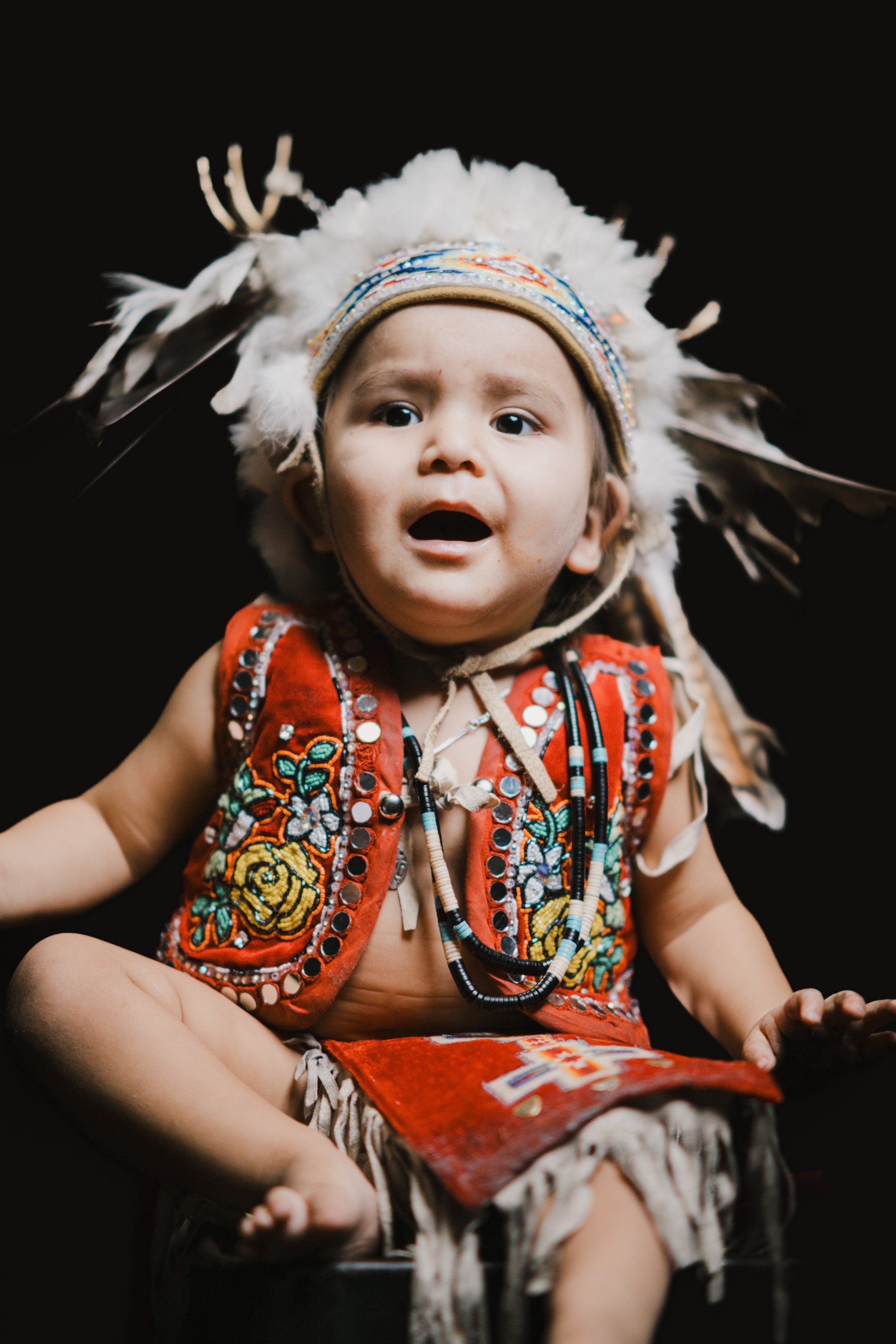 Young child in red Coast Salish regalia and white feathered headdress, mouth open in joy against dark background