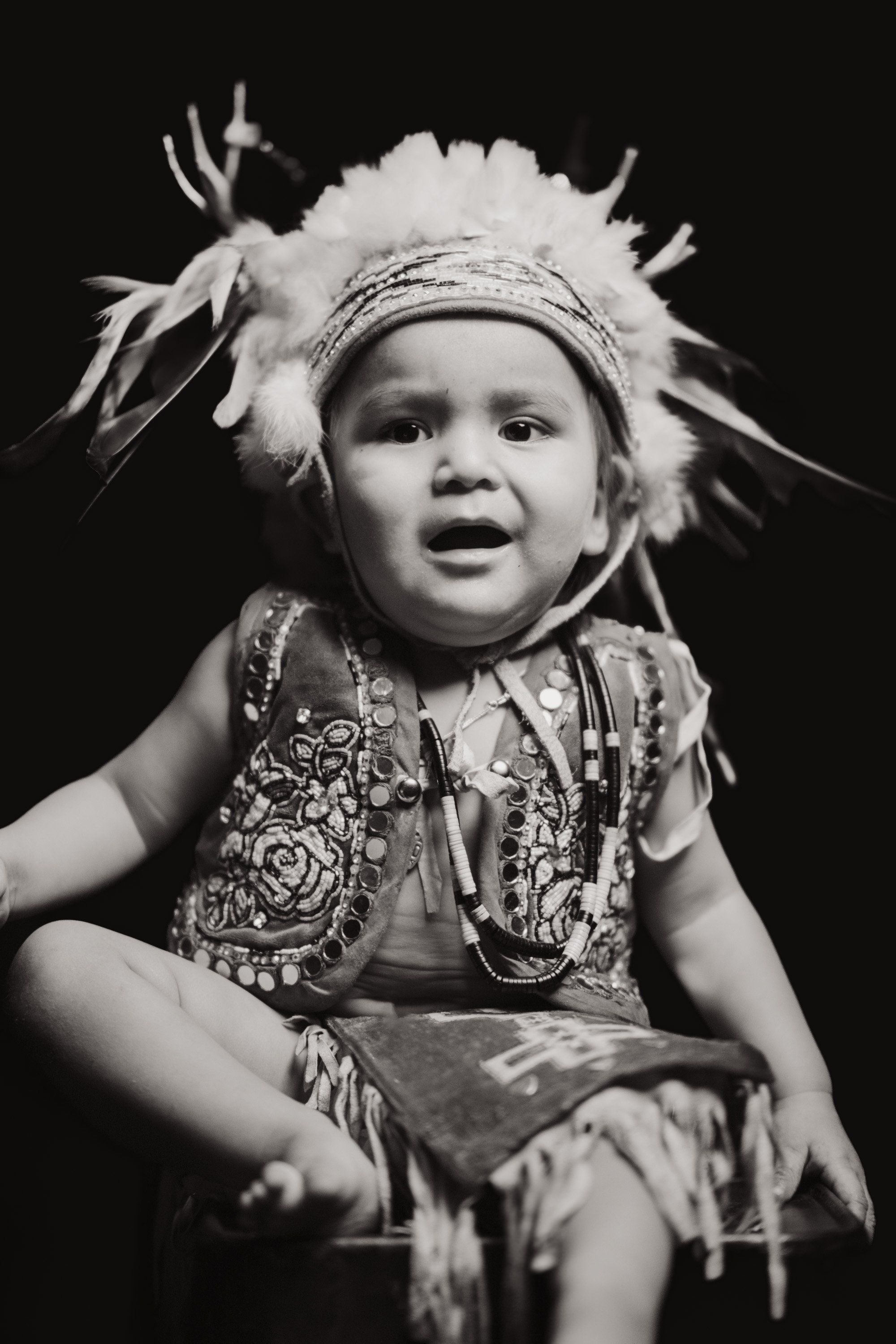 Young child in ornate regalia with fluffy headdress and beaded vest, mouth open in song against dark background