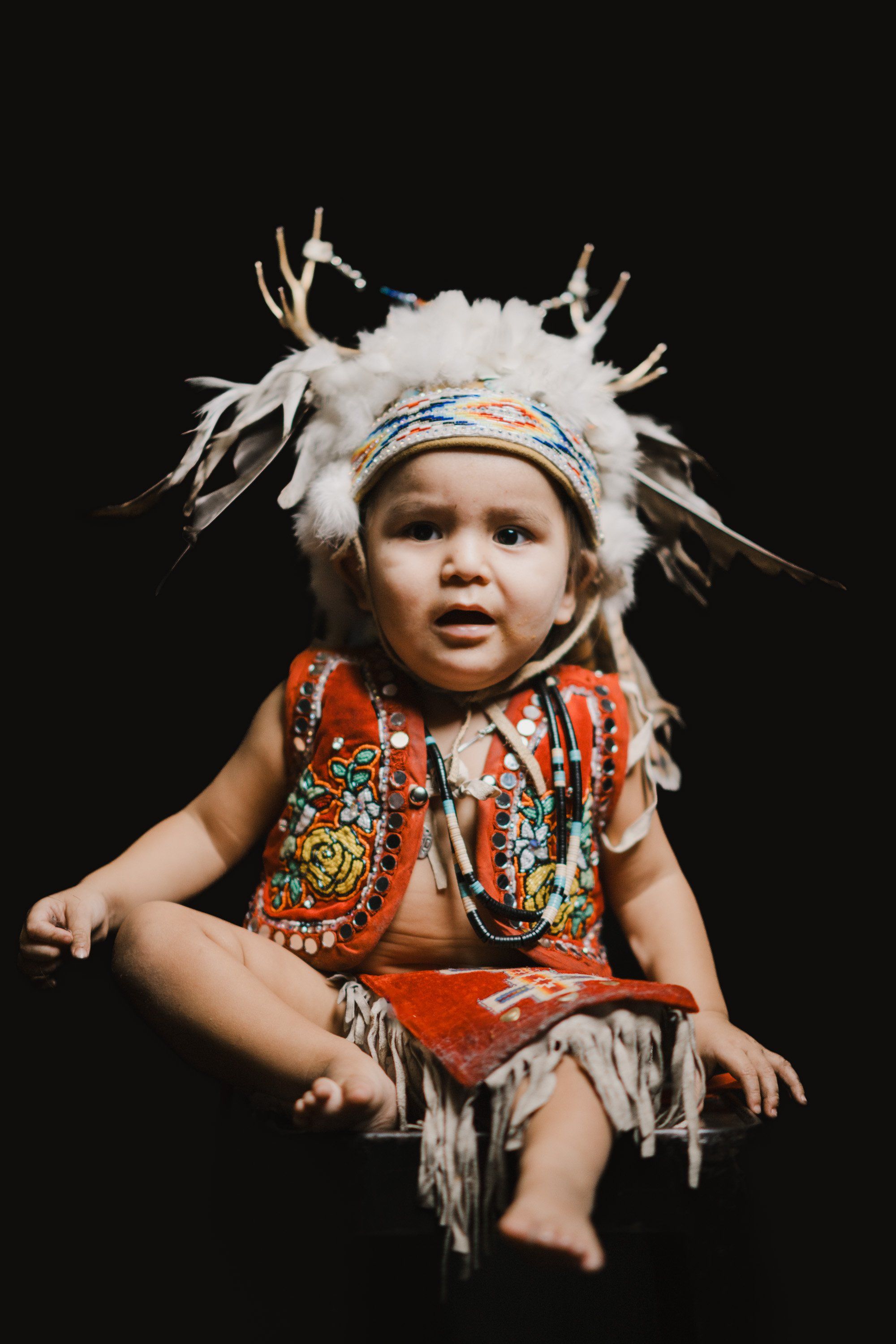 Toddler in traditional Coast Salish regalia with white fur headdress and red embroidered vest gazes upward against black background