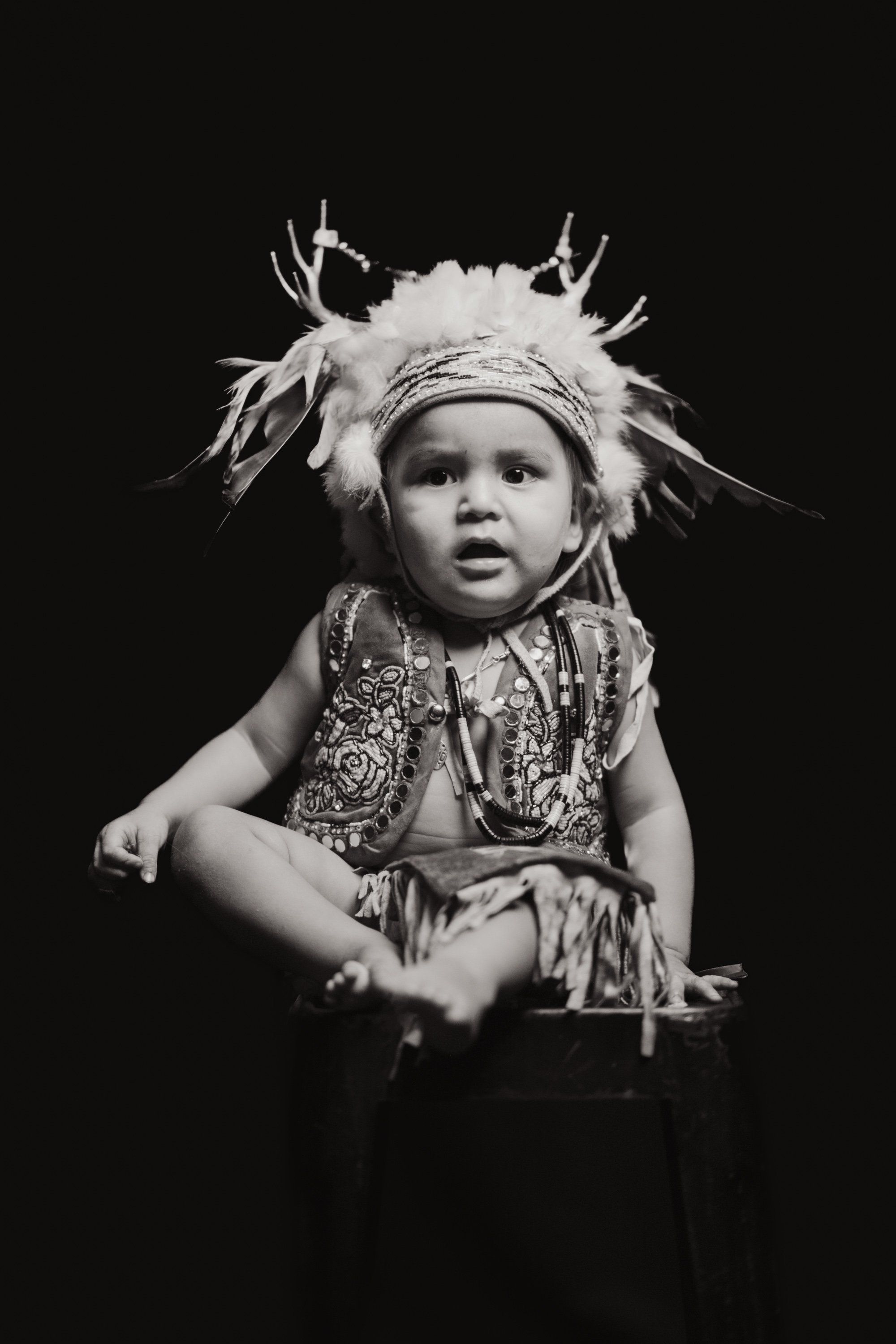 Young child in traditional Coast Salish regalia with ornate headpiece gazes upward against dark background