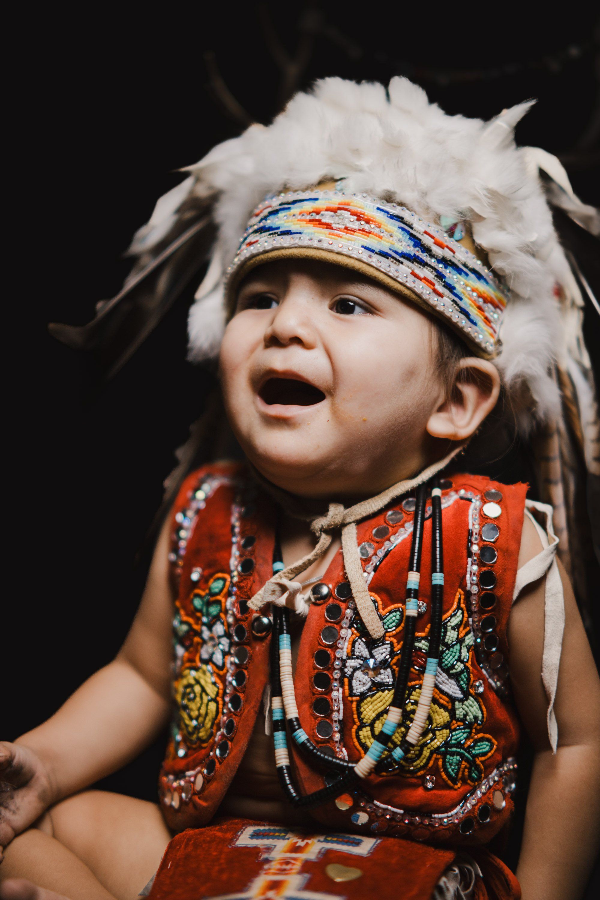 Young child in traditional Coast Salish regalia with white fur headdress and red button blanket, smiling with joy against dark background