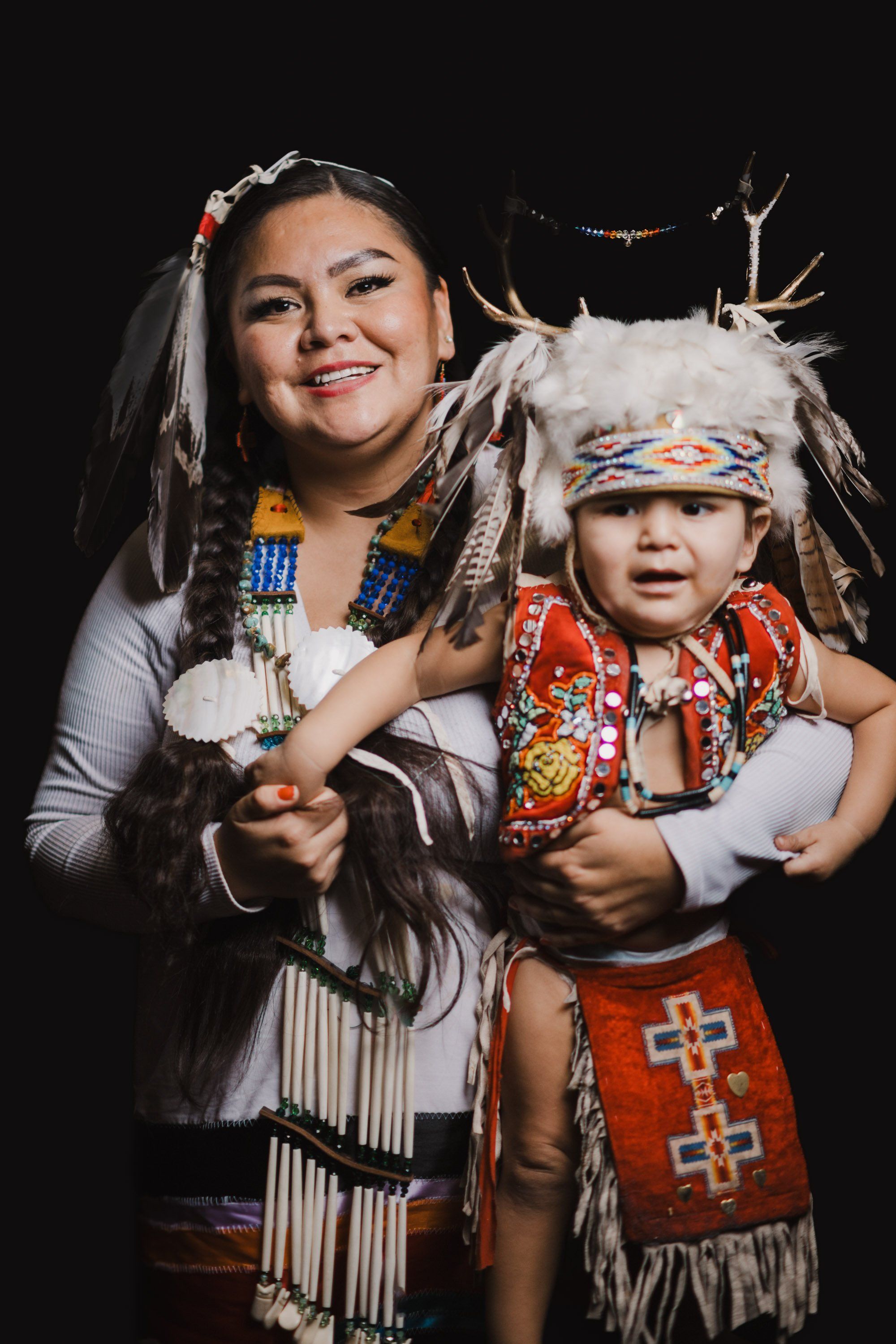 Woman and young child in traditional Coast Salish regalia with ornate beadwork and fringe against black background