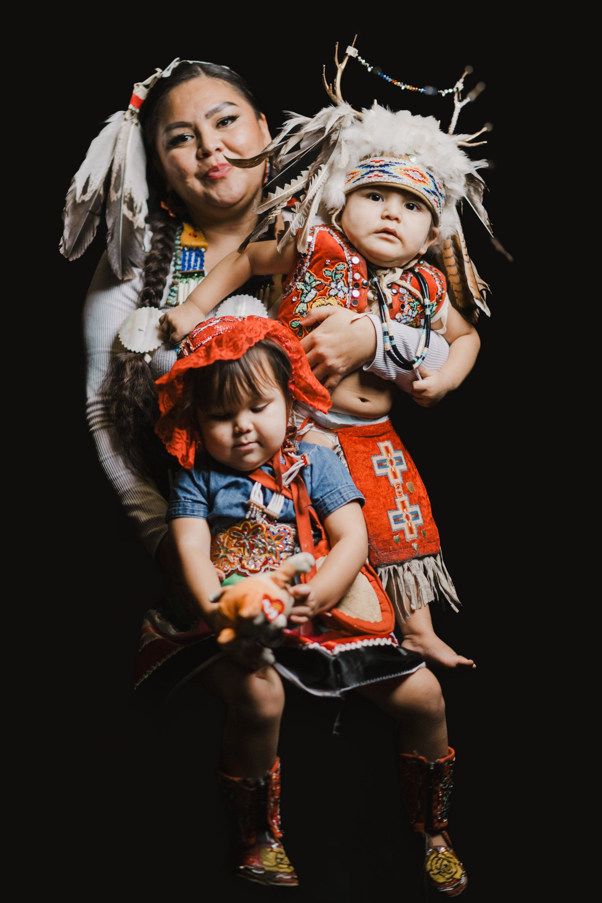 Woman in white-fringed regalia holds two young children in red and orange Coast Salish clothing against a black background