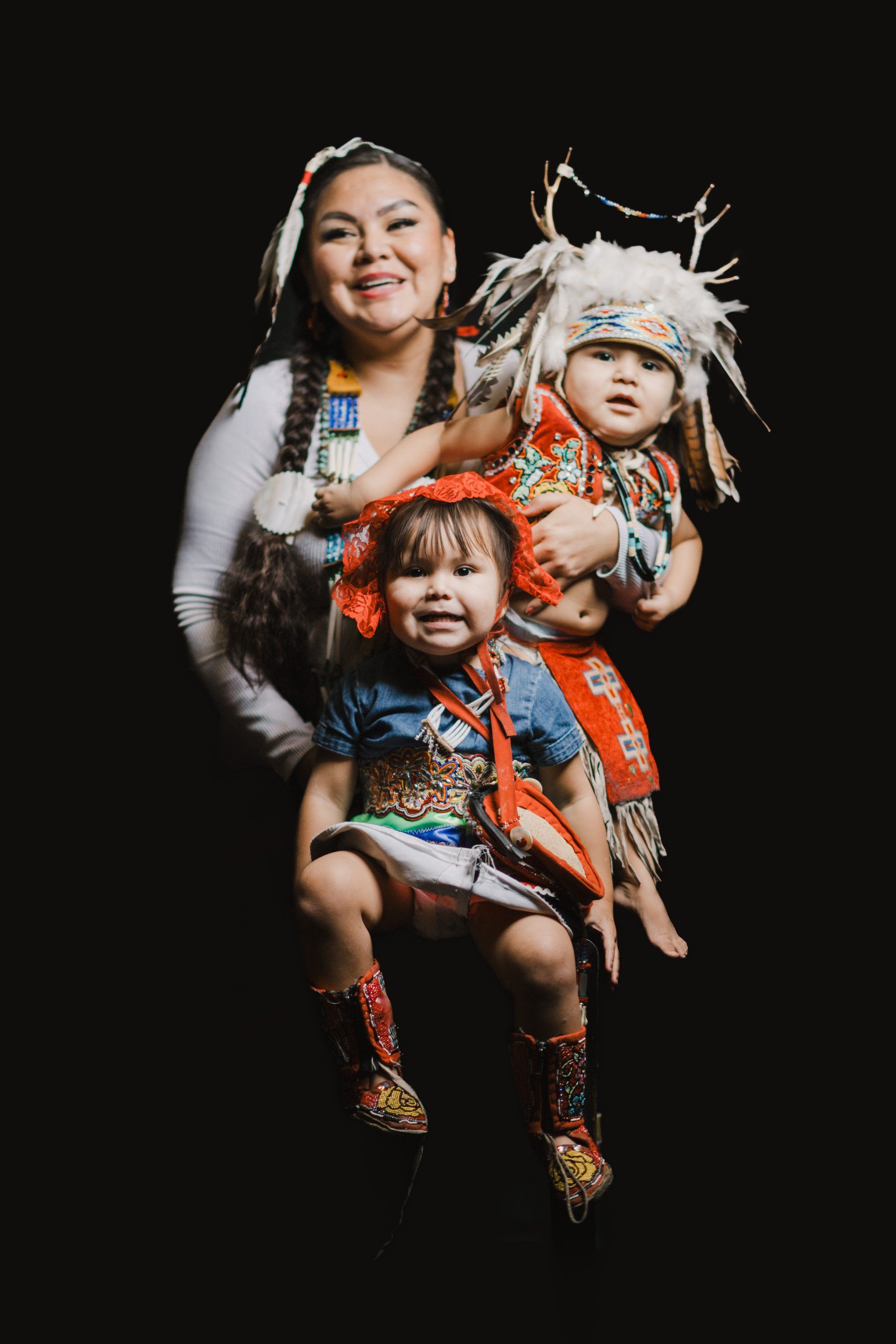Woman and two young children in Coast Salish regalia smiling against black background, holding traditional woven hats and blankets