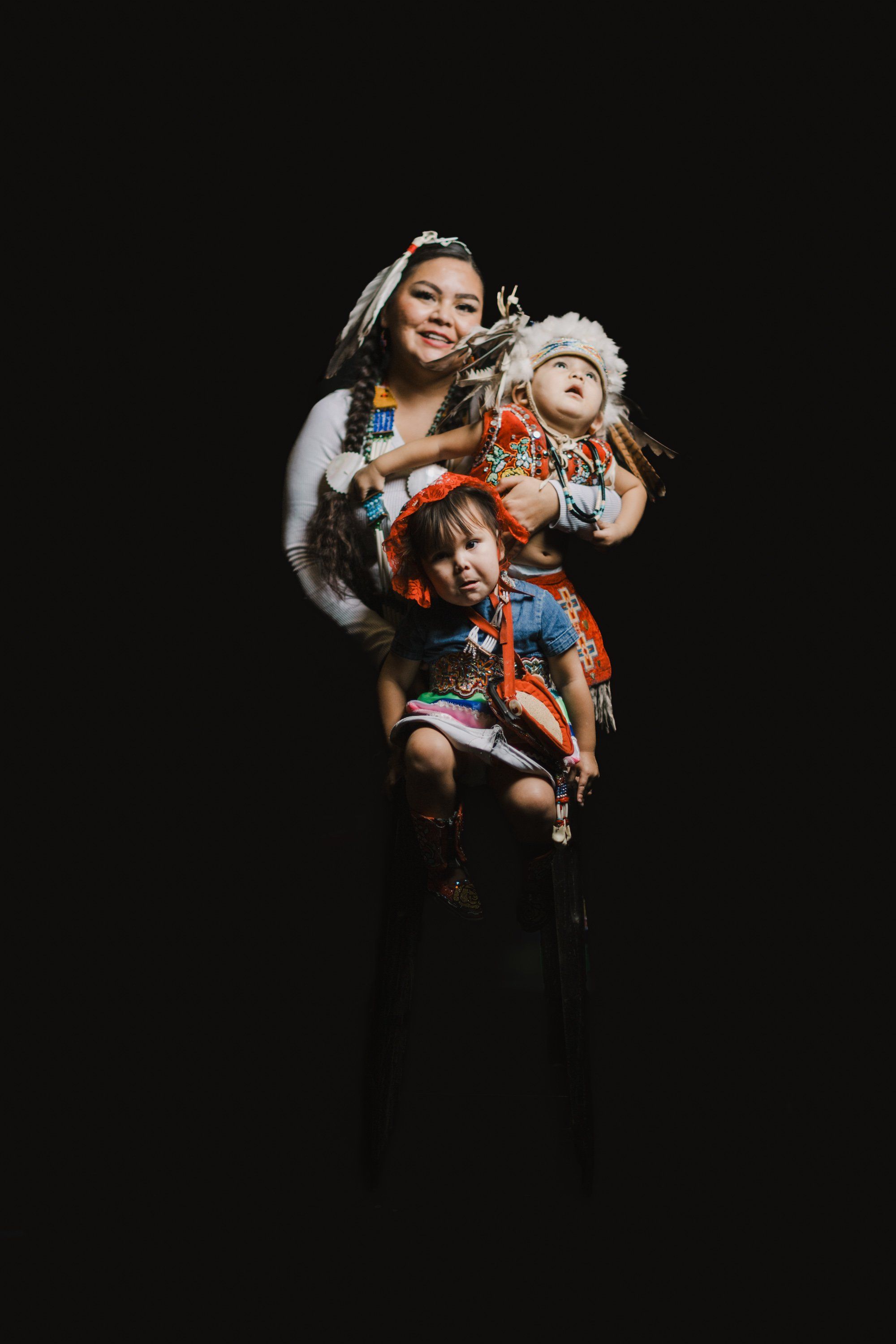 Woman in orange regalia holds a child and carved wolf mask against a black background, their expressions joyful and connected