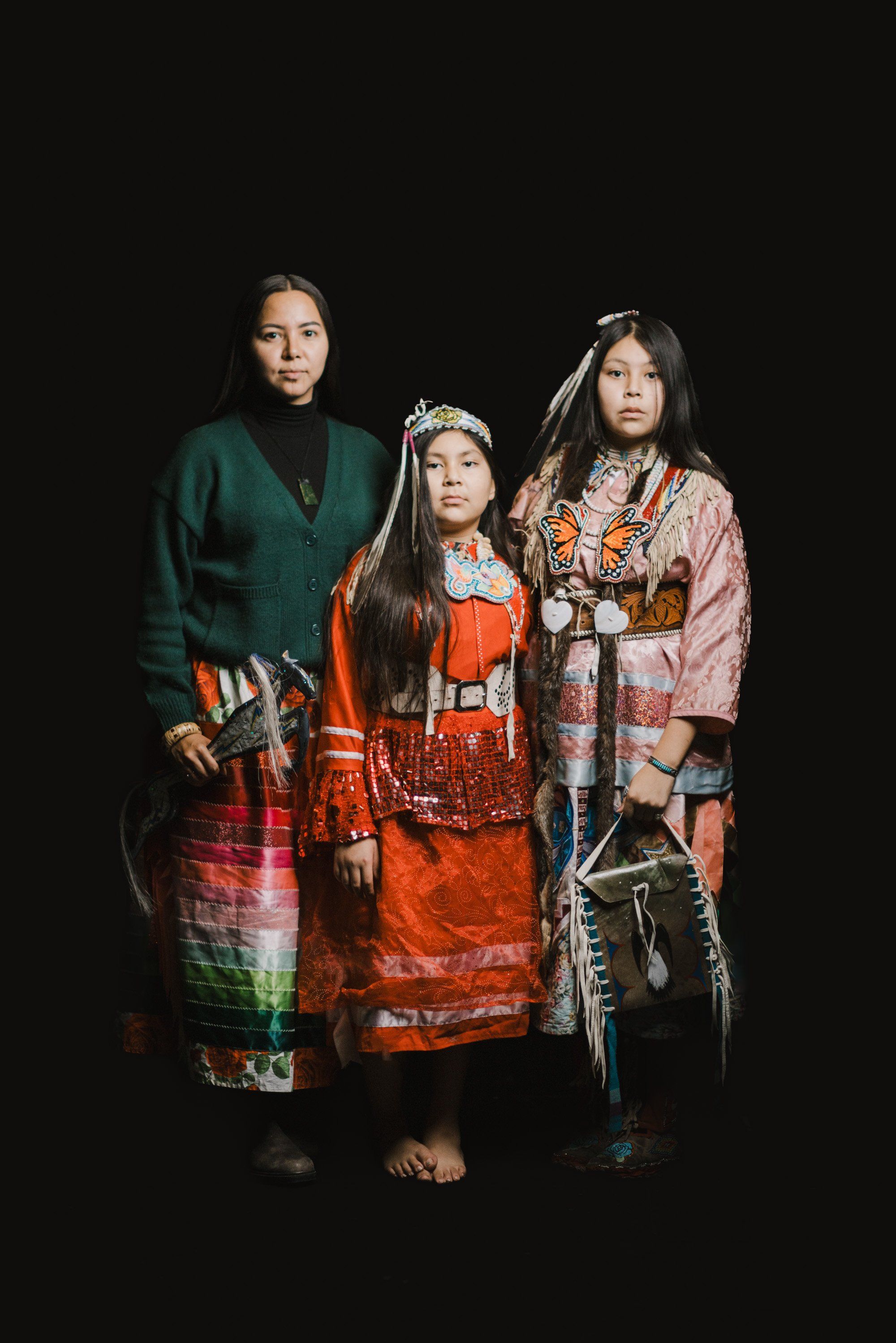 Three Coast Salish family members in traditional regalia stand against black background, wearing vibrant red, green, and pink woven blankets and beaded acces…
