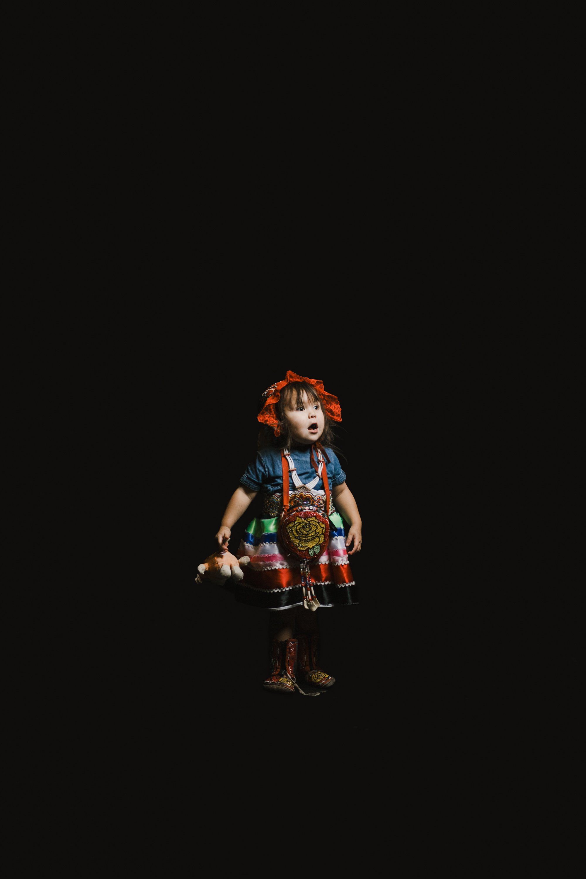 Young child in vibrant regalia with orange-red button blanket and headdress stands spotlit against black background, holding traditional dance objects