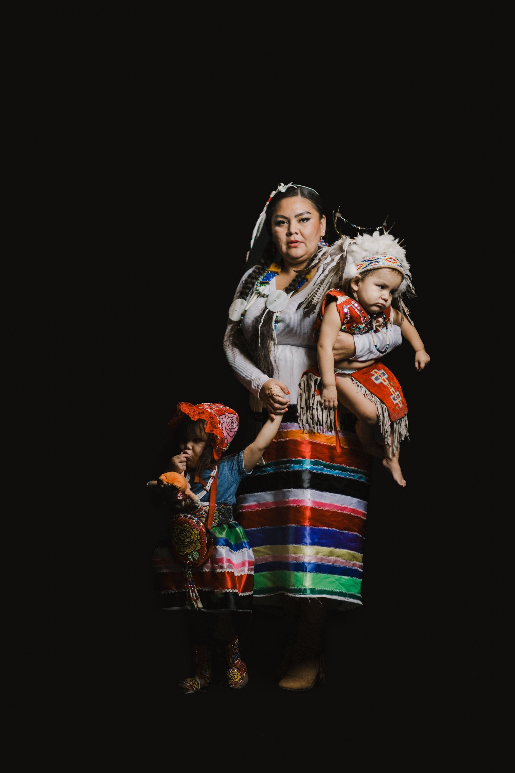 Woman in traditional Coast Salish regalia holding a child, both wearing blankets and ornate clothing, photographed against a black background