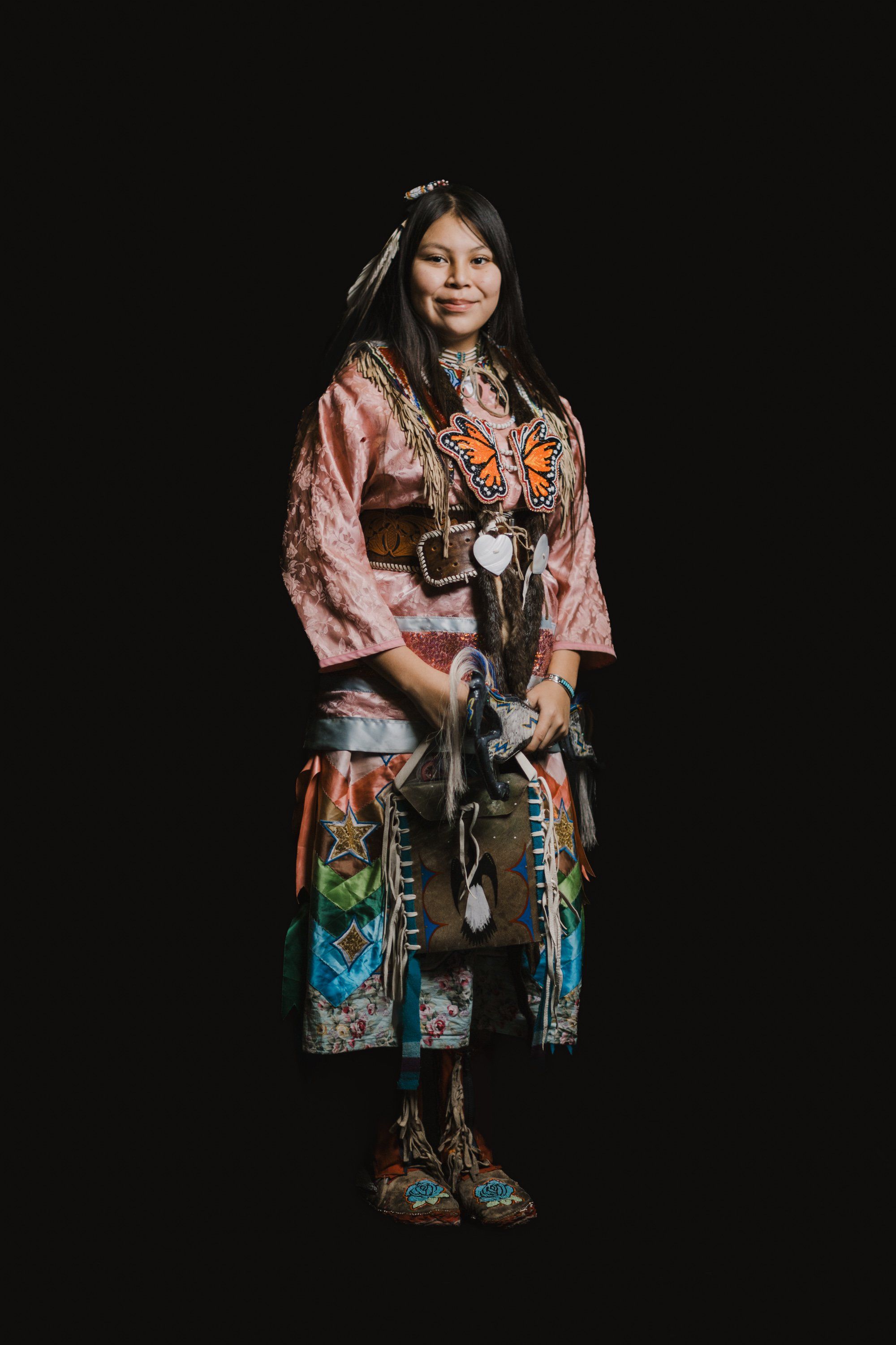 Young dancer in pink button-up shirt and embroidered regalia smiles at camera against black background, wearing traditional Coast Salish accessories and jewelry