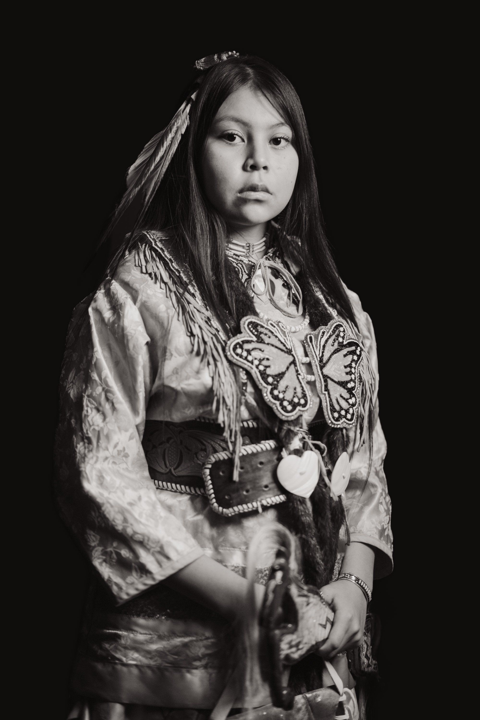 Young person in traditional Coast Salish regalia with butterfly design against dark background, direct gaze, dramatic studio lighting