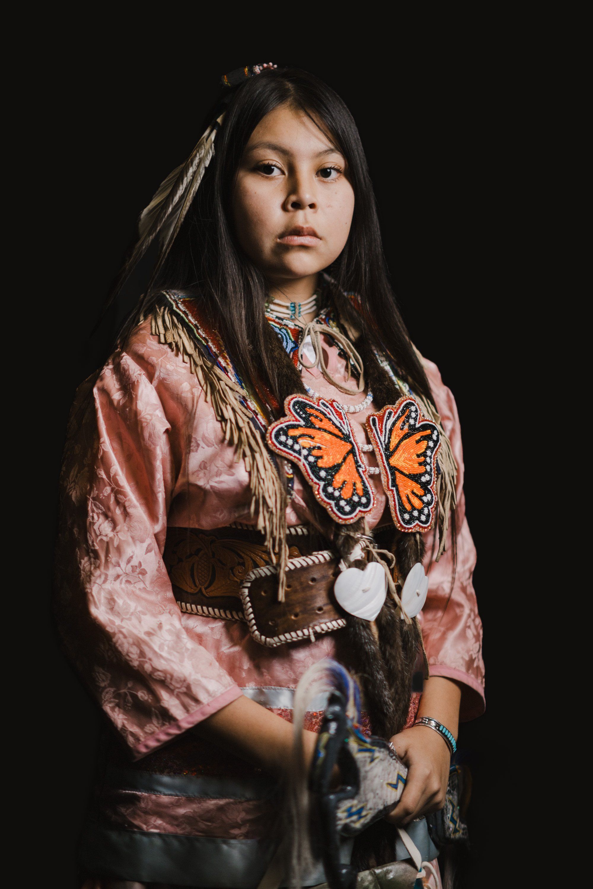 Young person in pink silk jacket with orange butterfly design, wearing traditional Coast Salish regalia and feathered hair ornaments, posed against dark back…