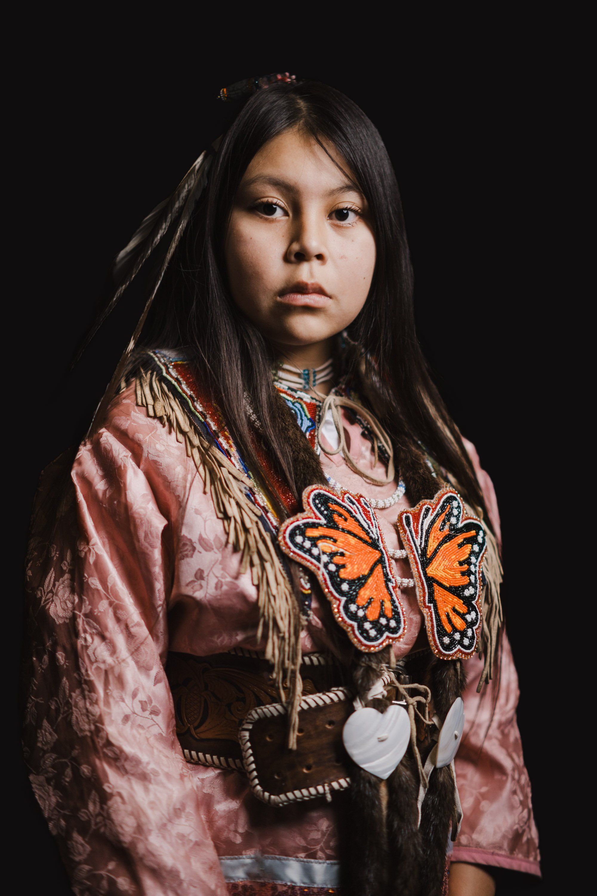 Young person in pink velvet regalia with orange butterfly design and fringe against black background, direct gaze, warm studio lighting