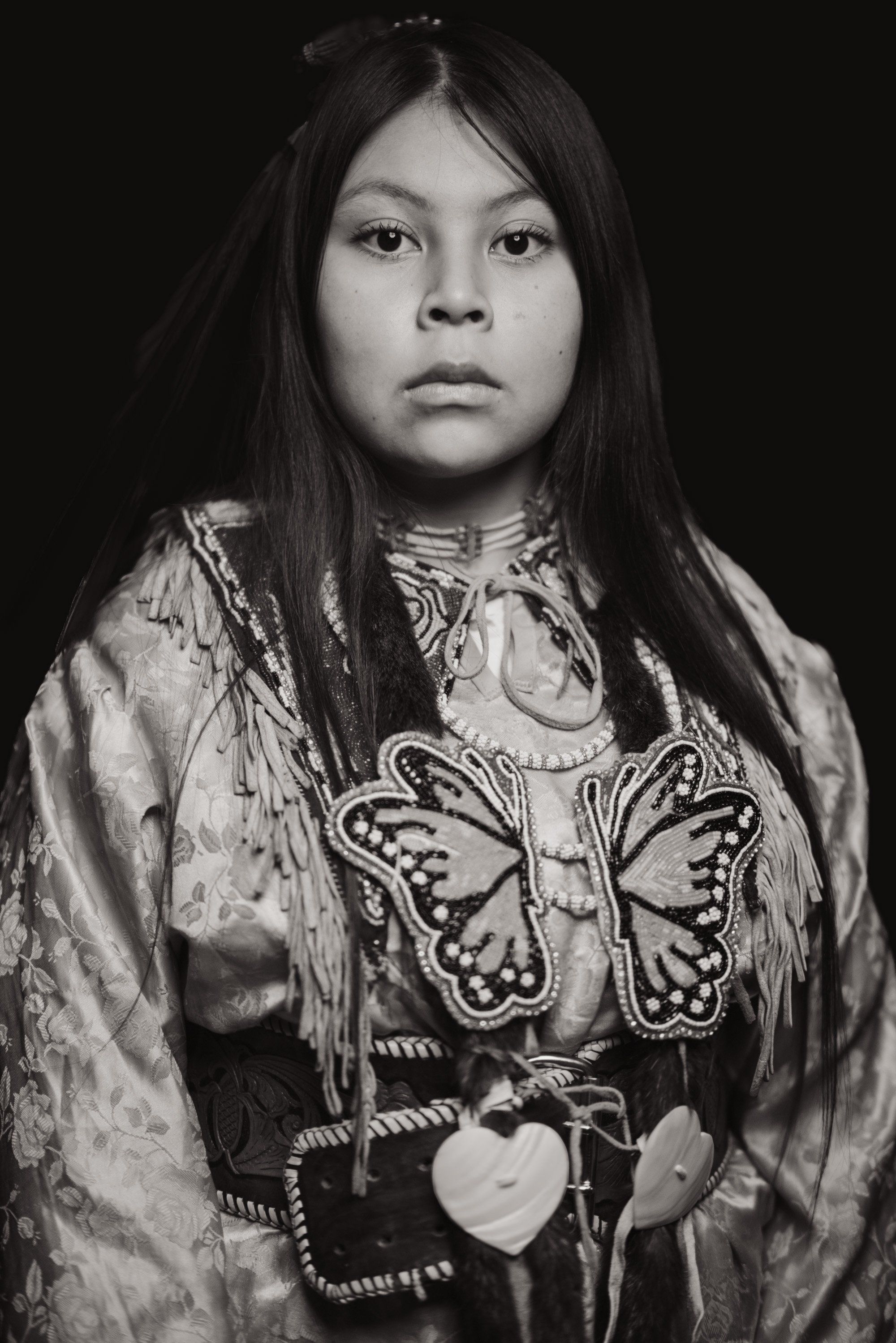 Young person in traditional Coast Salish regalia with butterfly design facing camera against black background, wearing woven tunic and pendant