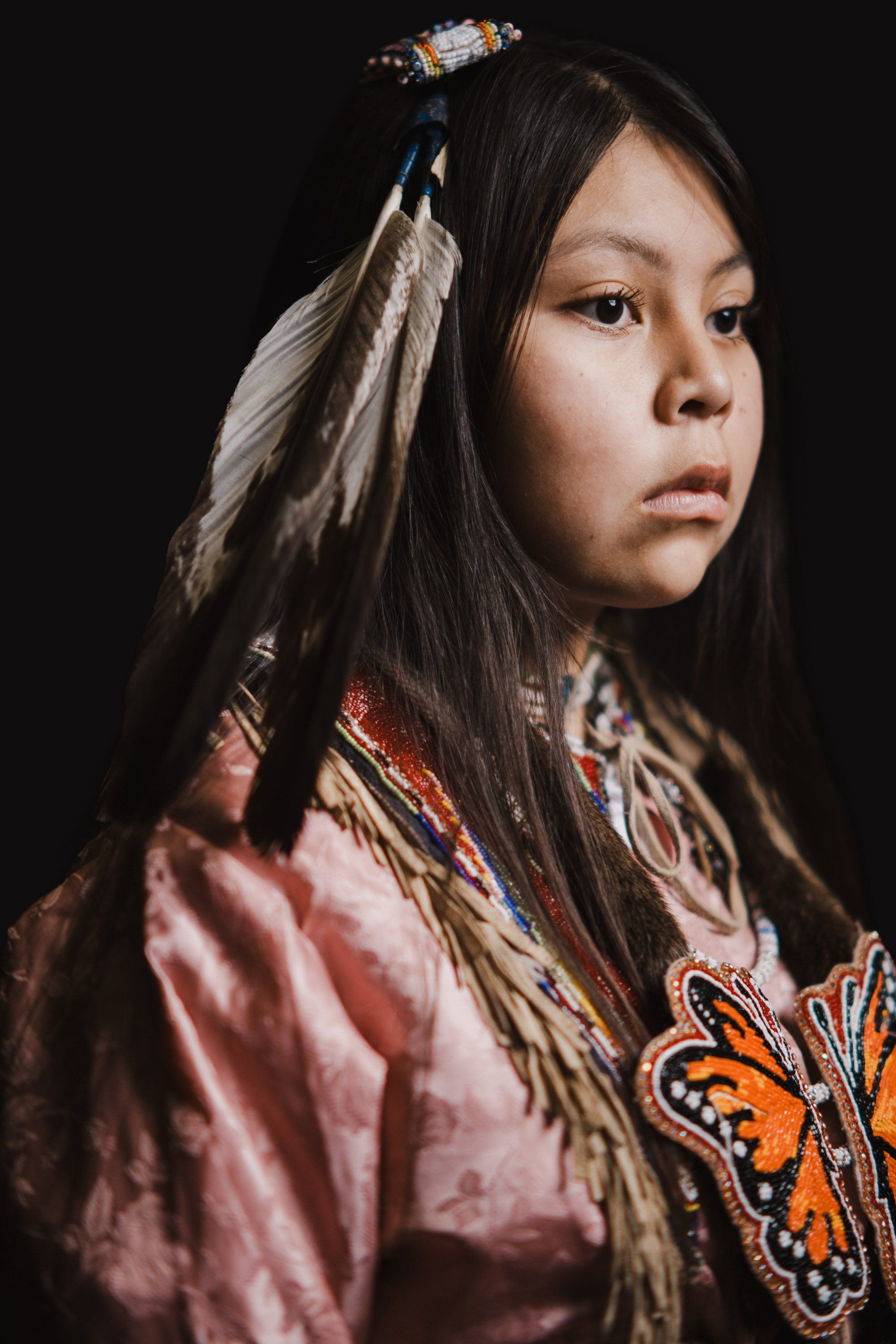 Young person in traditional Coast Salish regalia with feathered hair ornament, facing left against black background with warm studio lighting