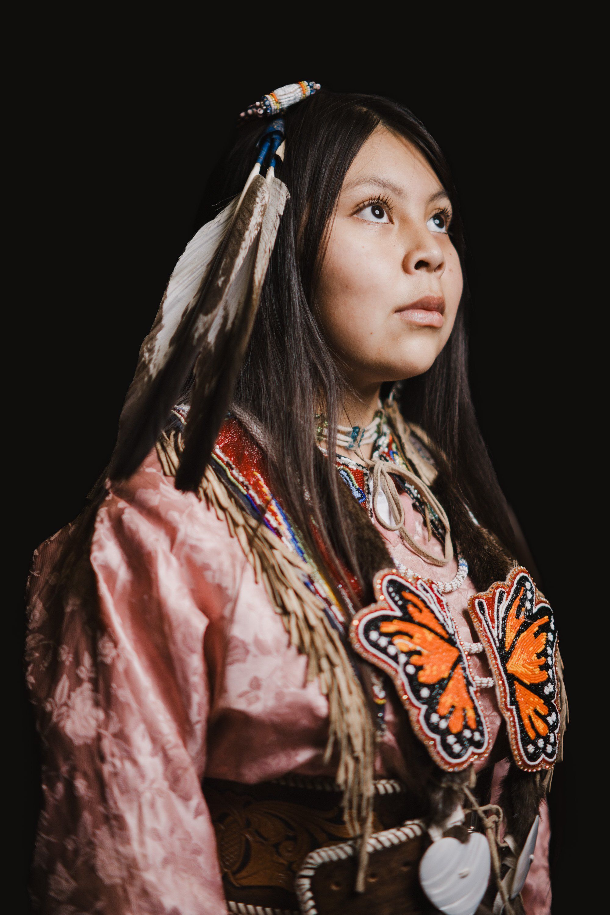 Young Coast Salish dancer in traditional regalia with butterfly design, beaded hair ornaments, and pink button blanket against black background