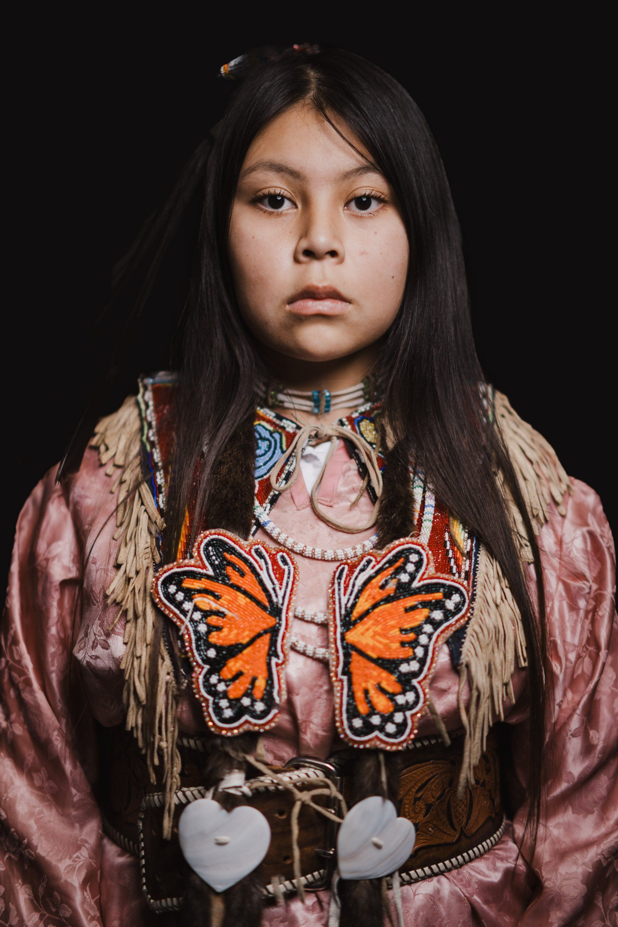 Young person in traditional Coast Salish regalia with embroidered butterfly motifs and shell ornaments, dark background, dignified expression