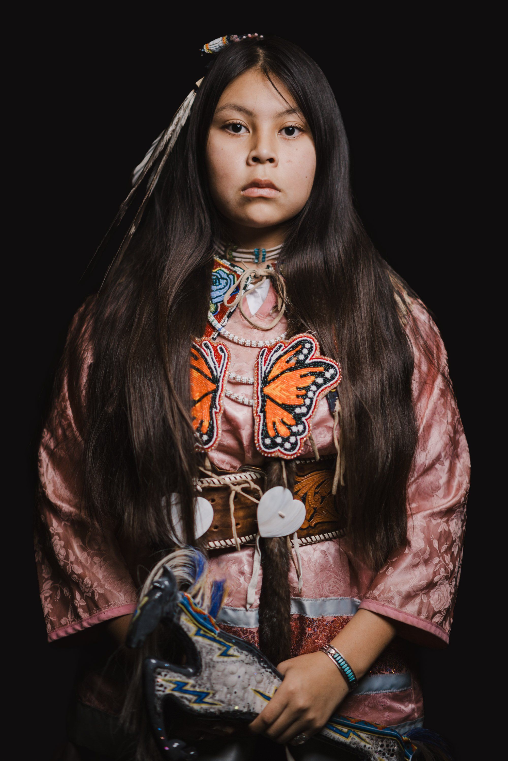 Young person with long black hair wearing a pink silk jacket and beaded regalia featuring butterfly and geometric designs, photographed against black background