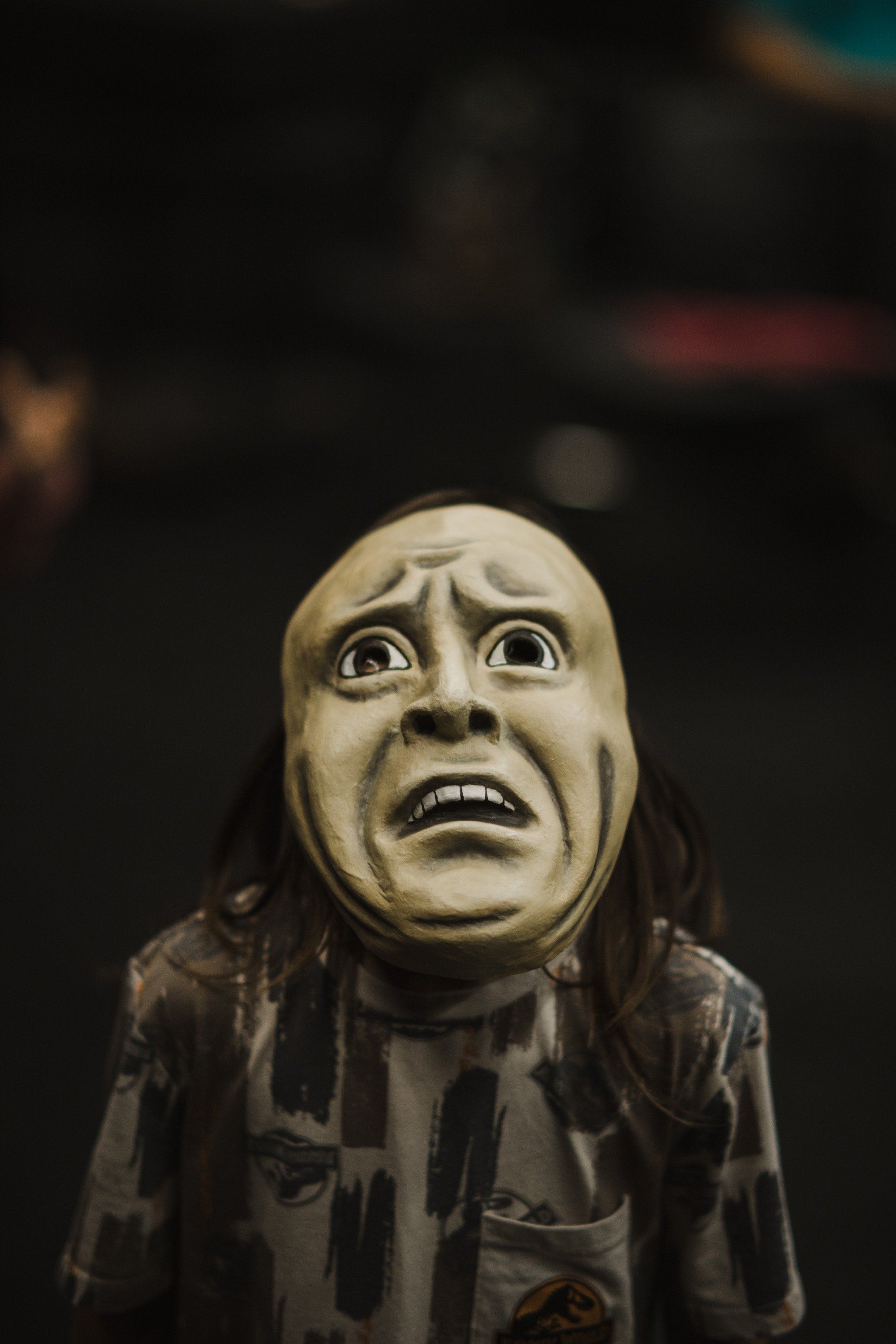 Carved wooden mask with wide eyes and open-mouthed expression, draped in dark patterned fabric against a blurred dark background