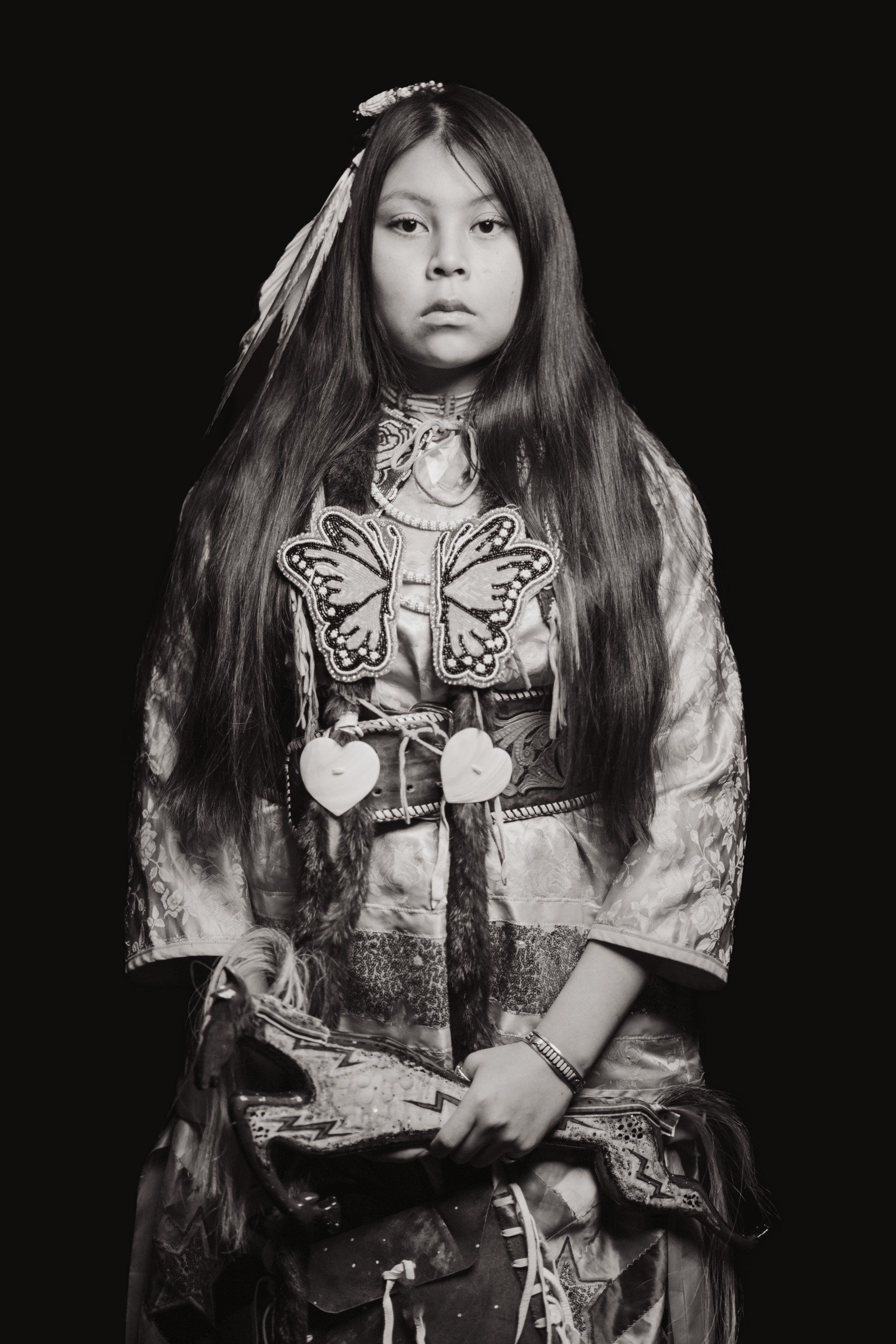 Young person with long dark hair adorned with white feathers, wearing ornate button blanket regalia with butterfly motif against black background