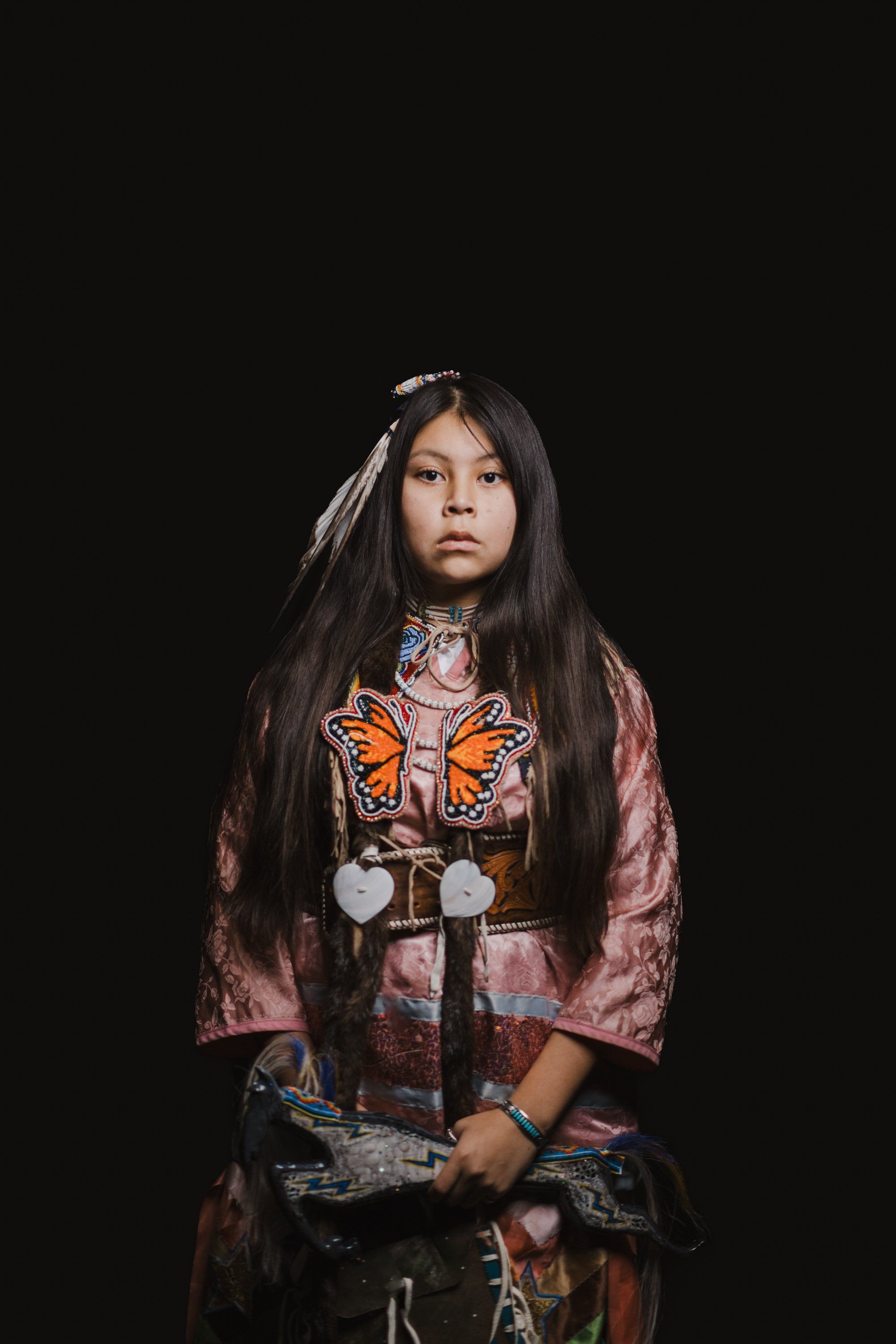 Young person in traditional Coast Salish regalia with orange butterfly design facing camera against black background, long dark hair with accessories, contem…
