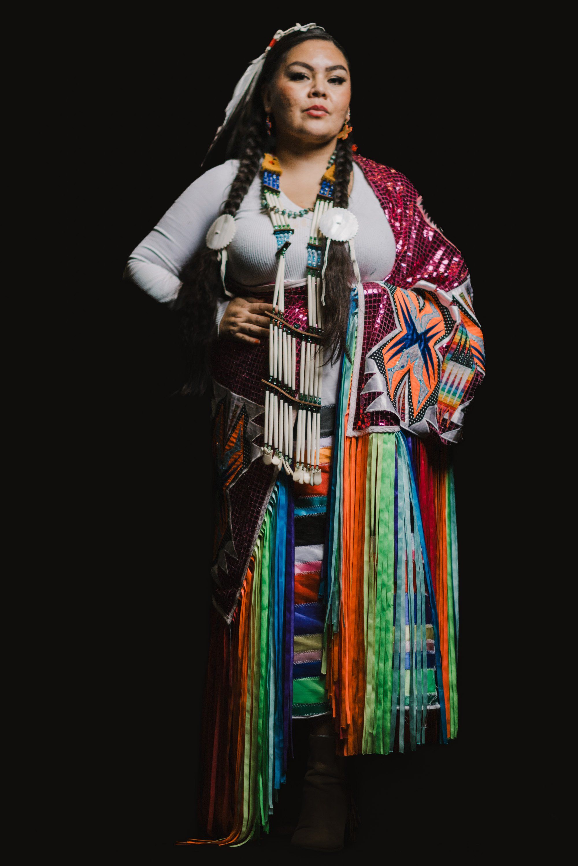 Coast Salish woman in traditional regalia with colorful fringed shawl and beaded accessories stands against black background
