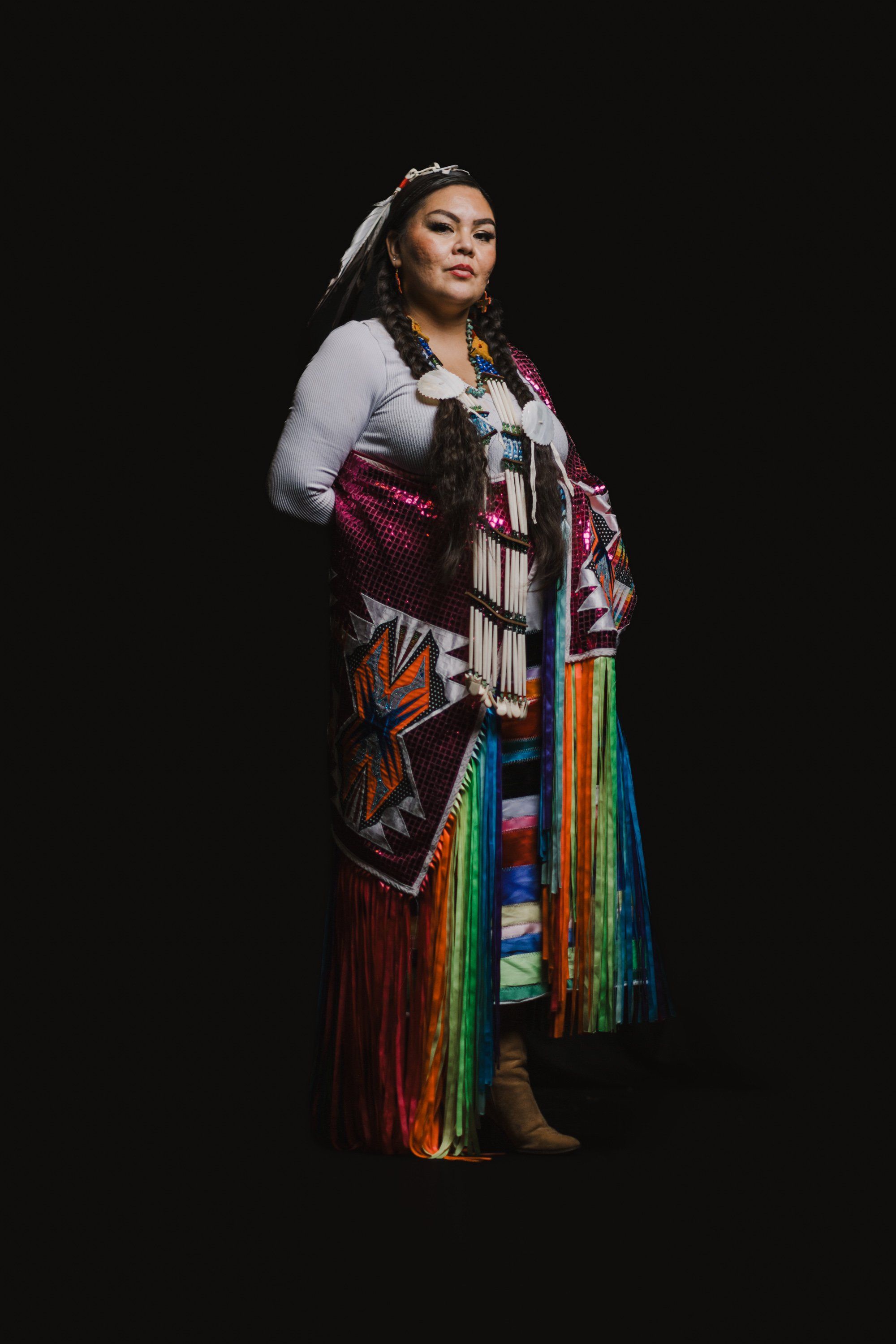 Woman in traditional Coast Salish regalia with red button blanket and colorful woven skirt stands against black background, looking upward