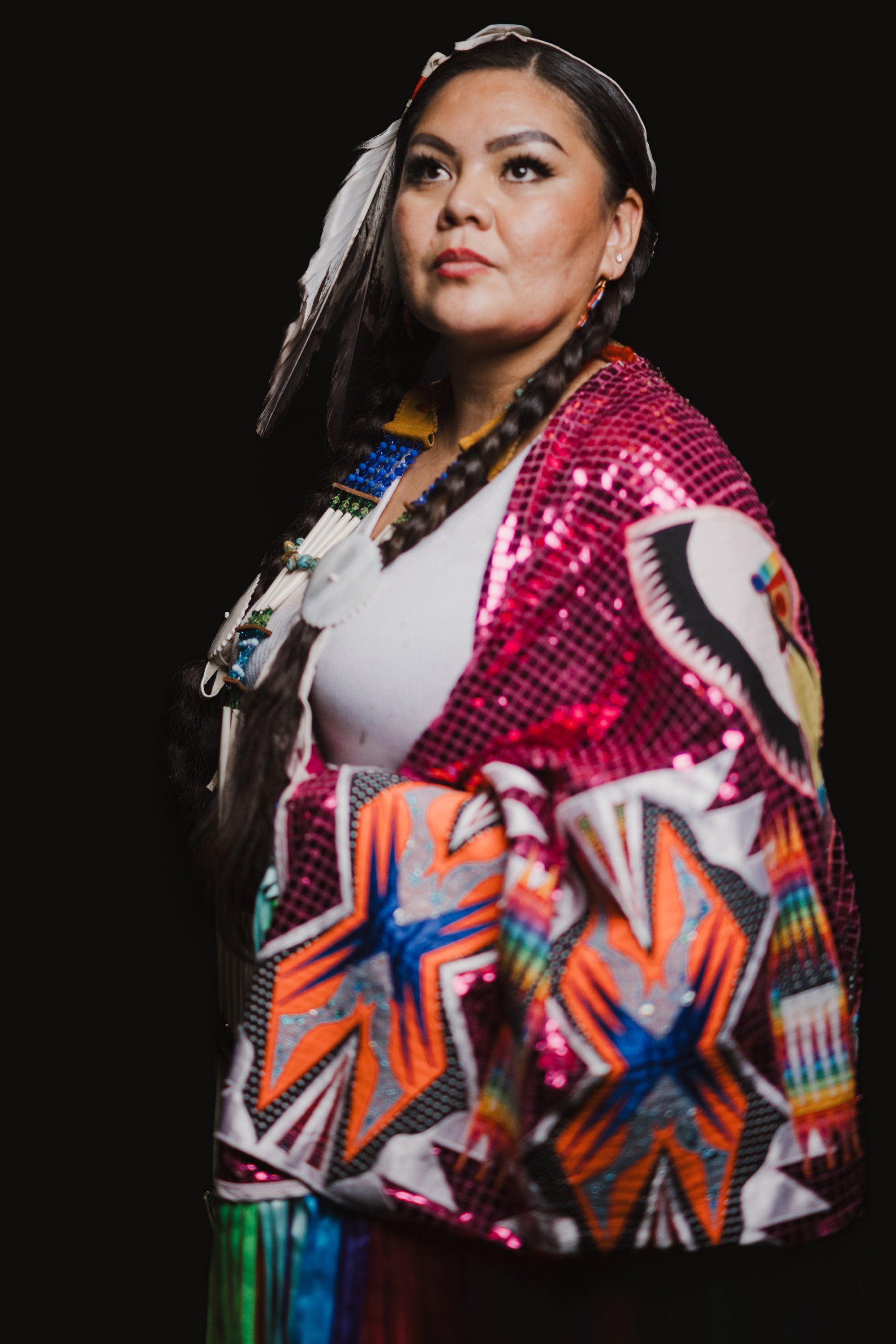 Woman in vibrant pink and orange patterned jacket gazes upward against black background, wearing white top and traditional regalia details