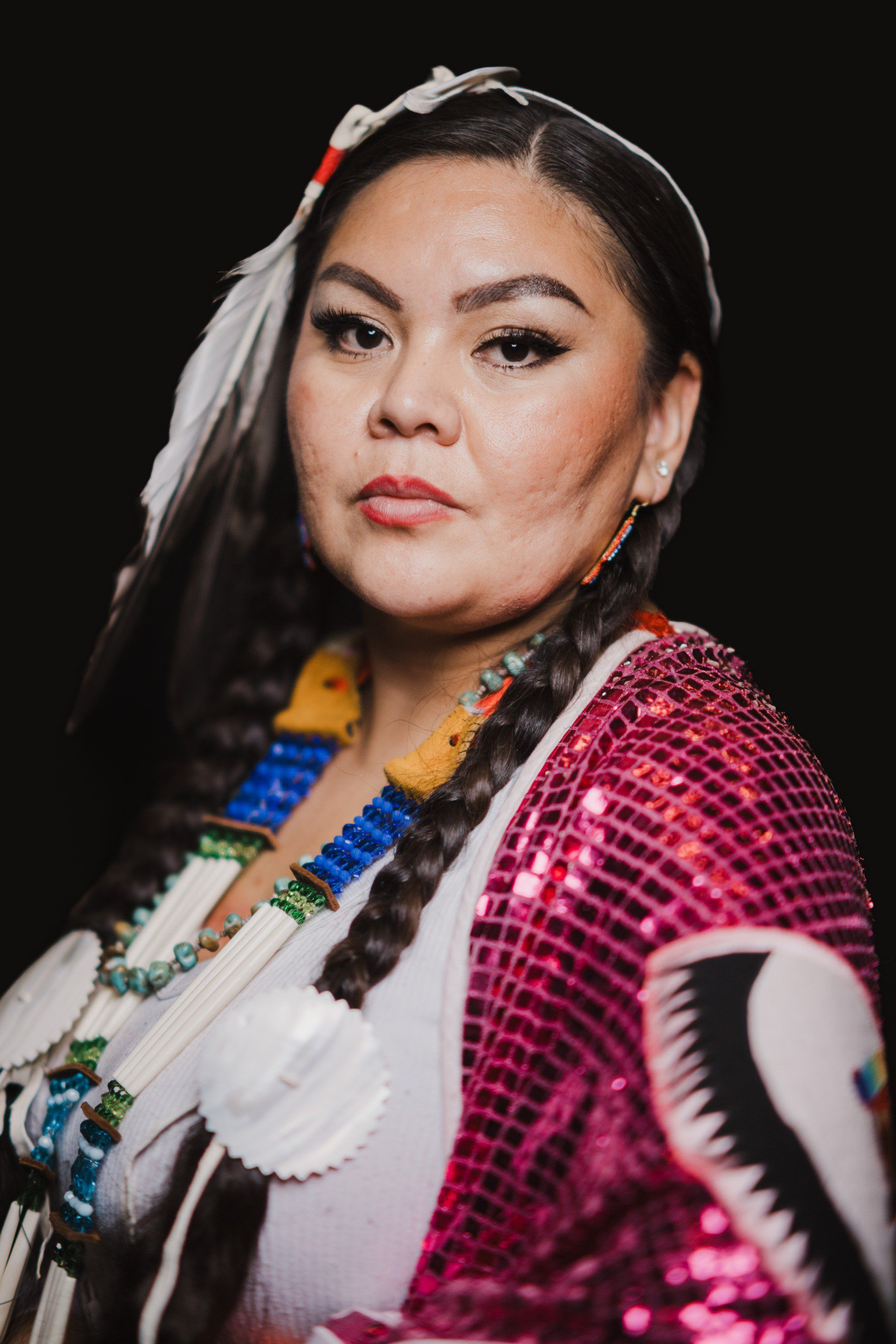 Woman in vibrant fuchsia regalia with white collar and beaded necklace gazes toward camera against black background