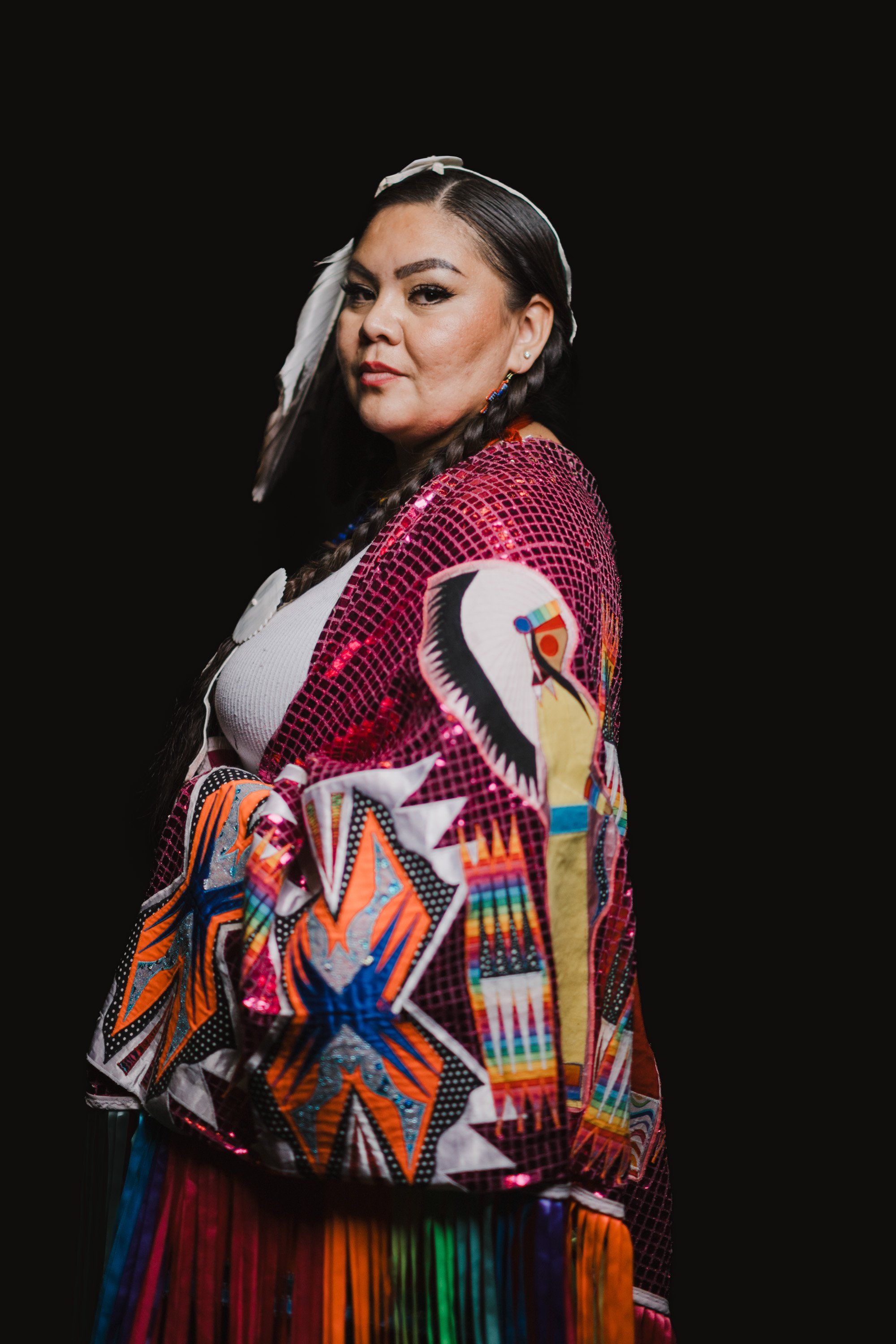 Woman in traditional Coast Salish regalia with burgundy button blanket and crane design turns toward camera against black background