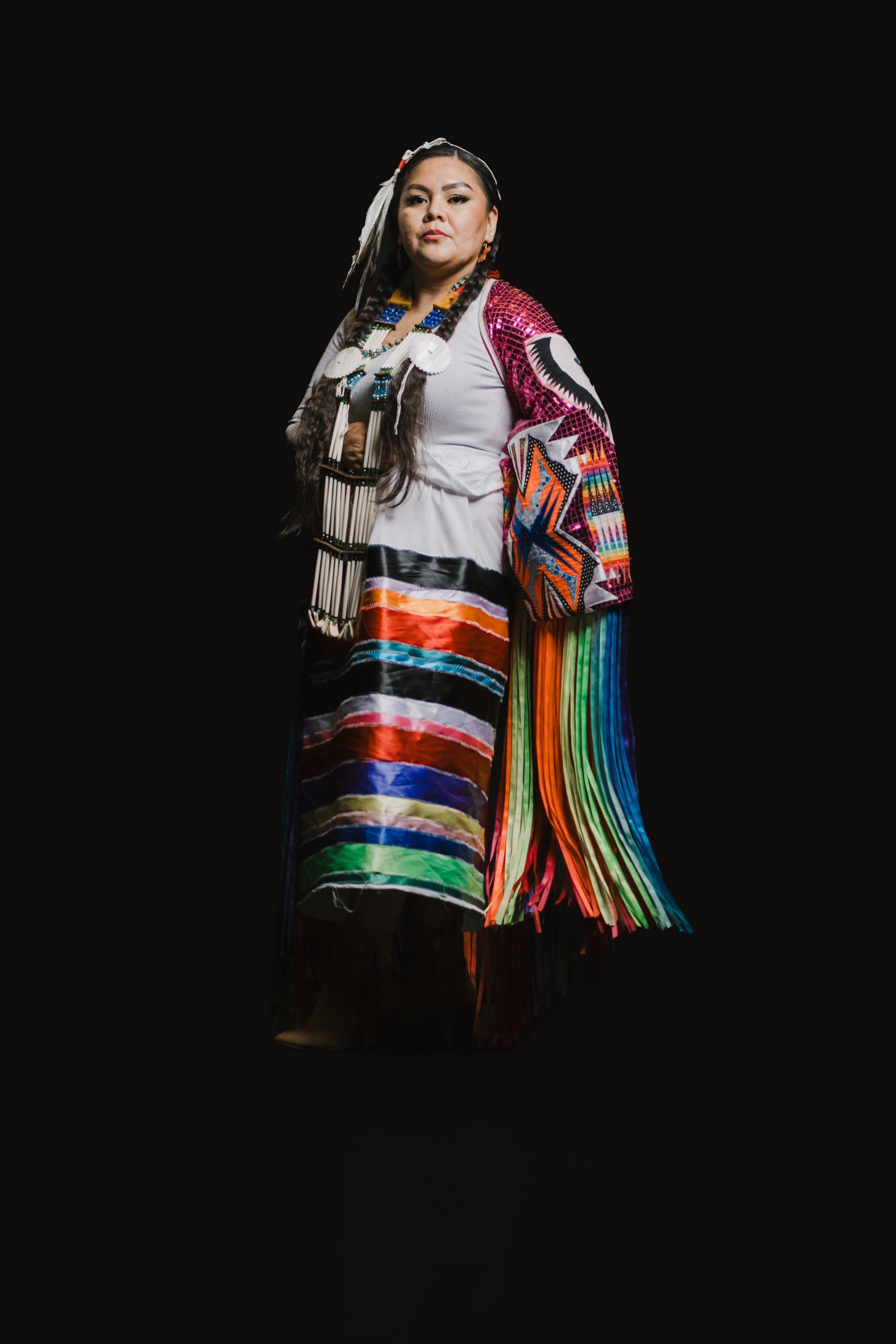 Person in traditional Coast Salish regalia with colorful striped blanket and white fringe shawl stands against black background, gazing upward