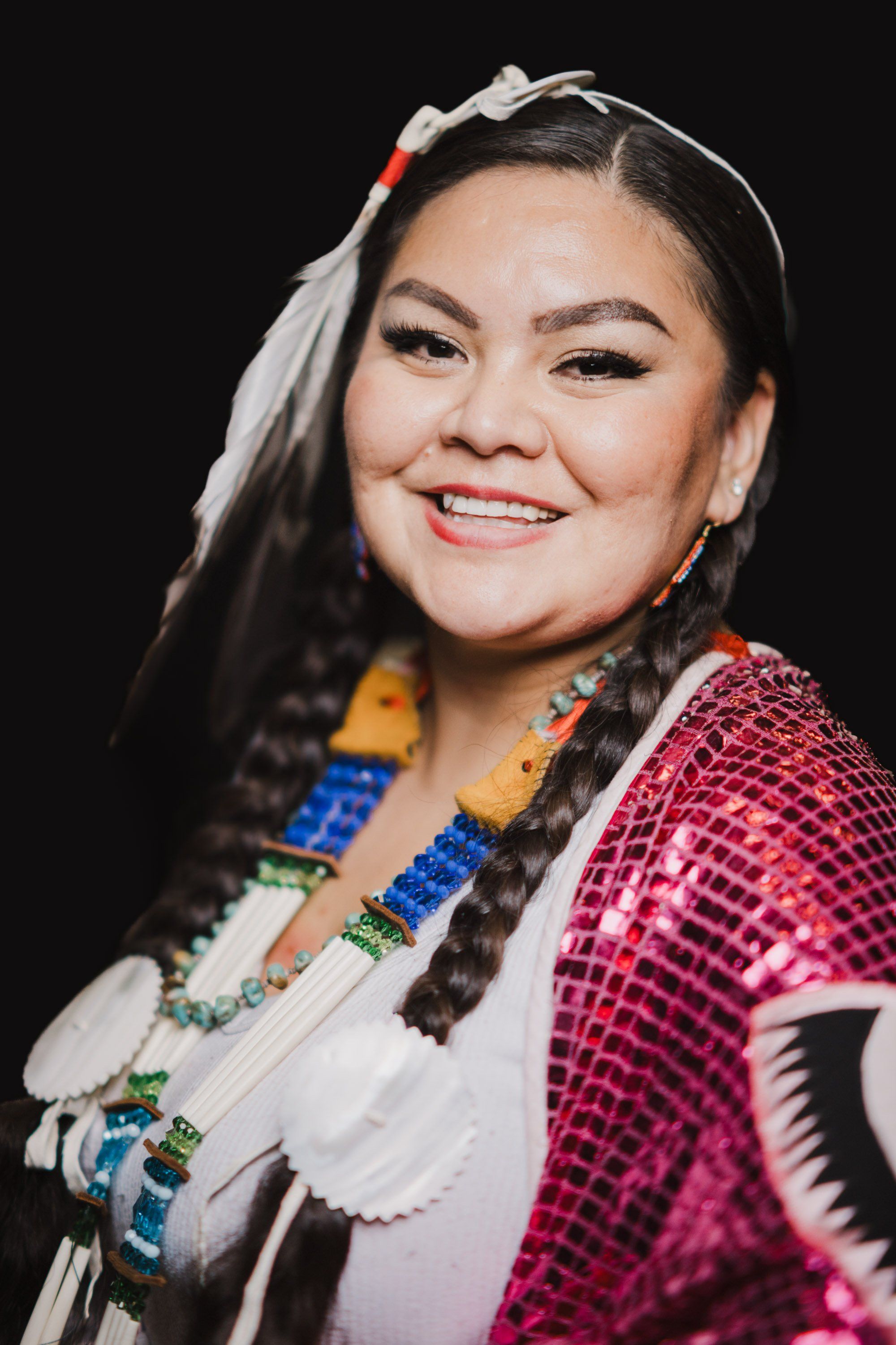Smiling person with long braided hair adorned with red beads, wearing a vibrant pink sequined shirt and traditional Coast Salish regalia against a black back…