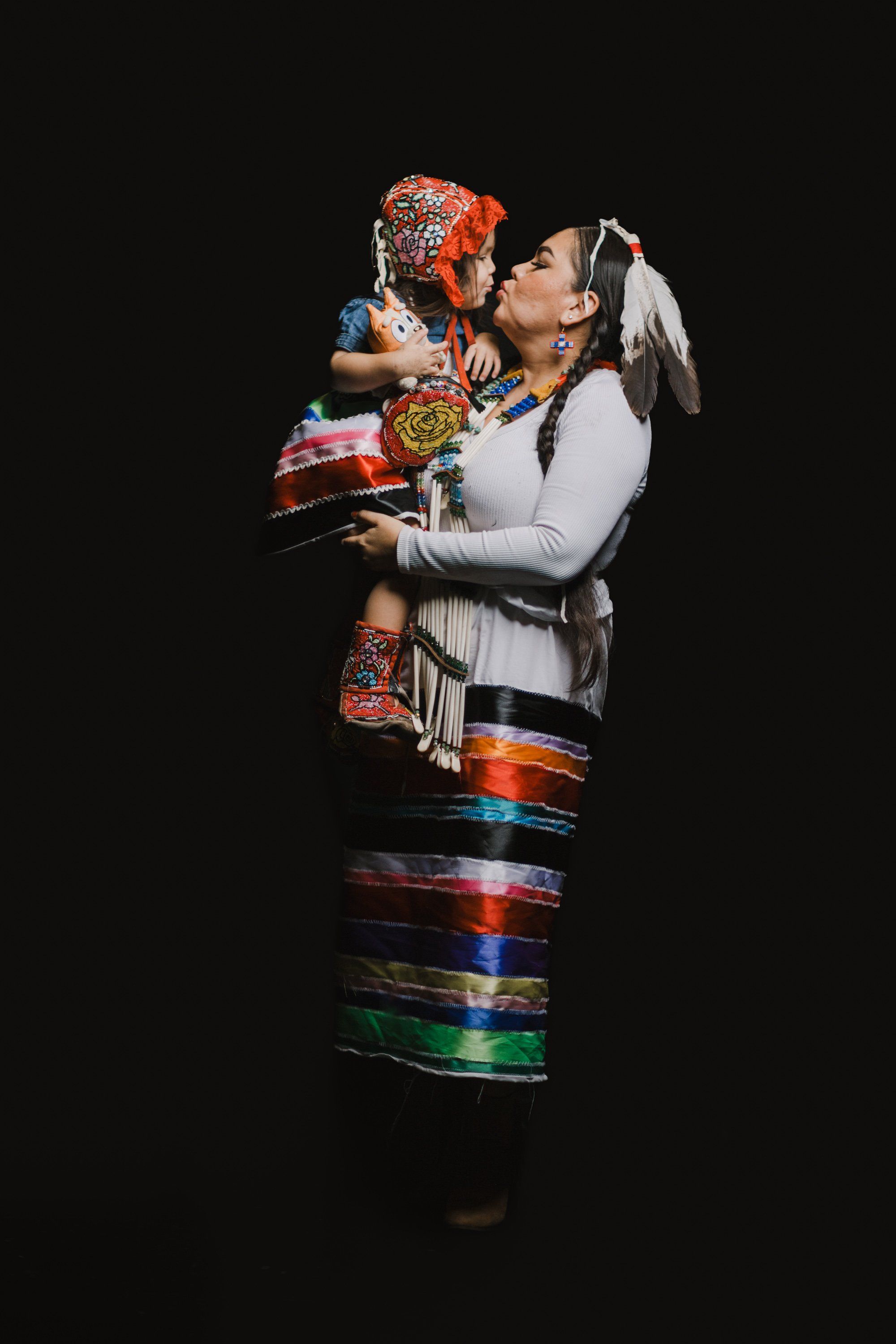 Woman in white regalia holding a child, both wearing vibrant Coast Salish textiles against black background