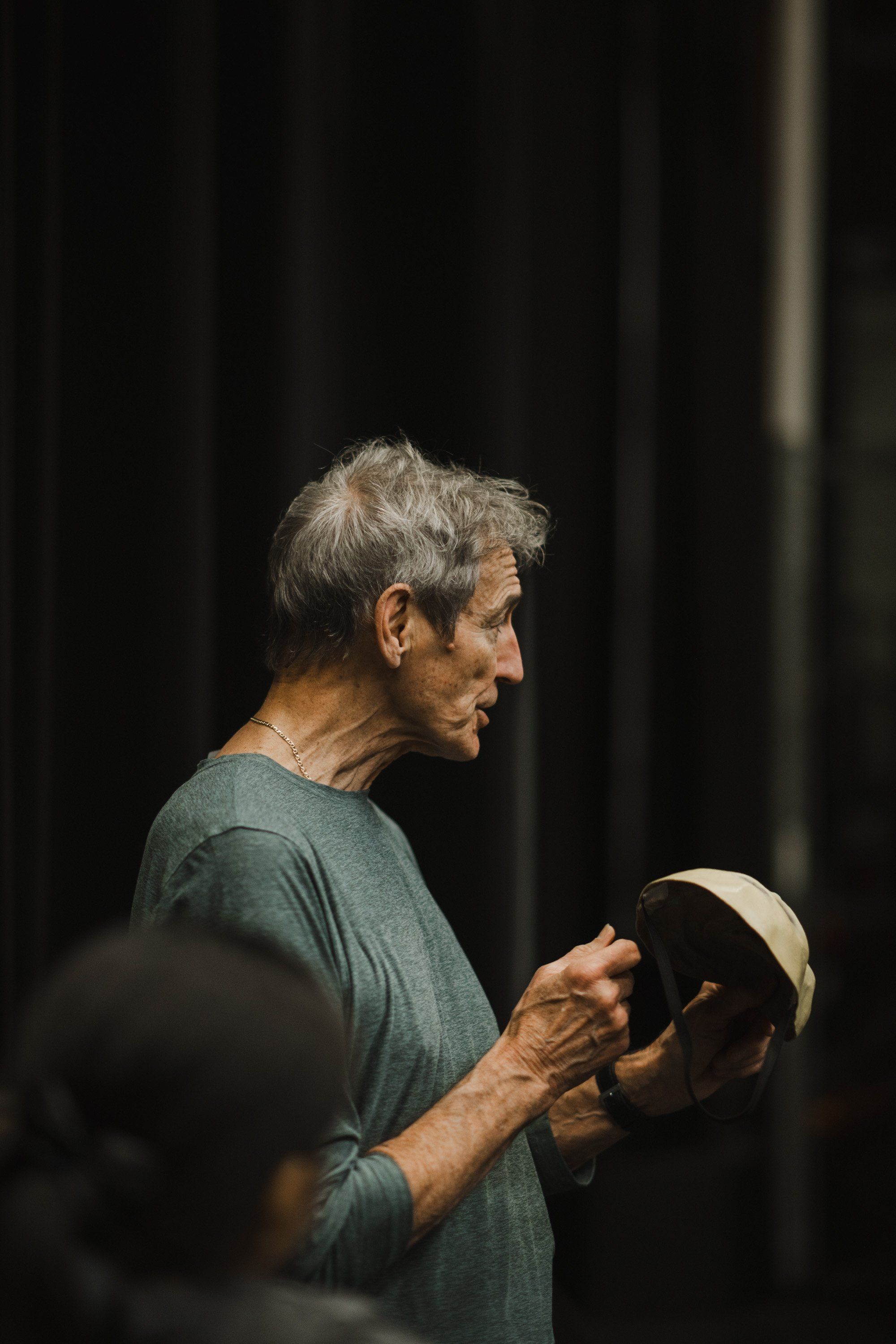 Elder in profile holding a traditional drum, speaking to an engaged audience against a dark backdrop