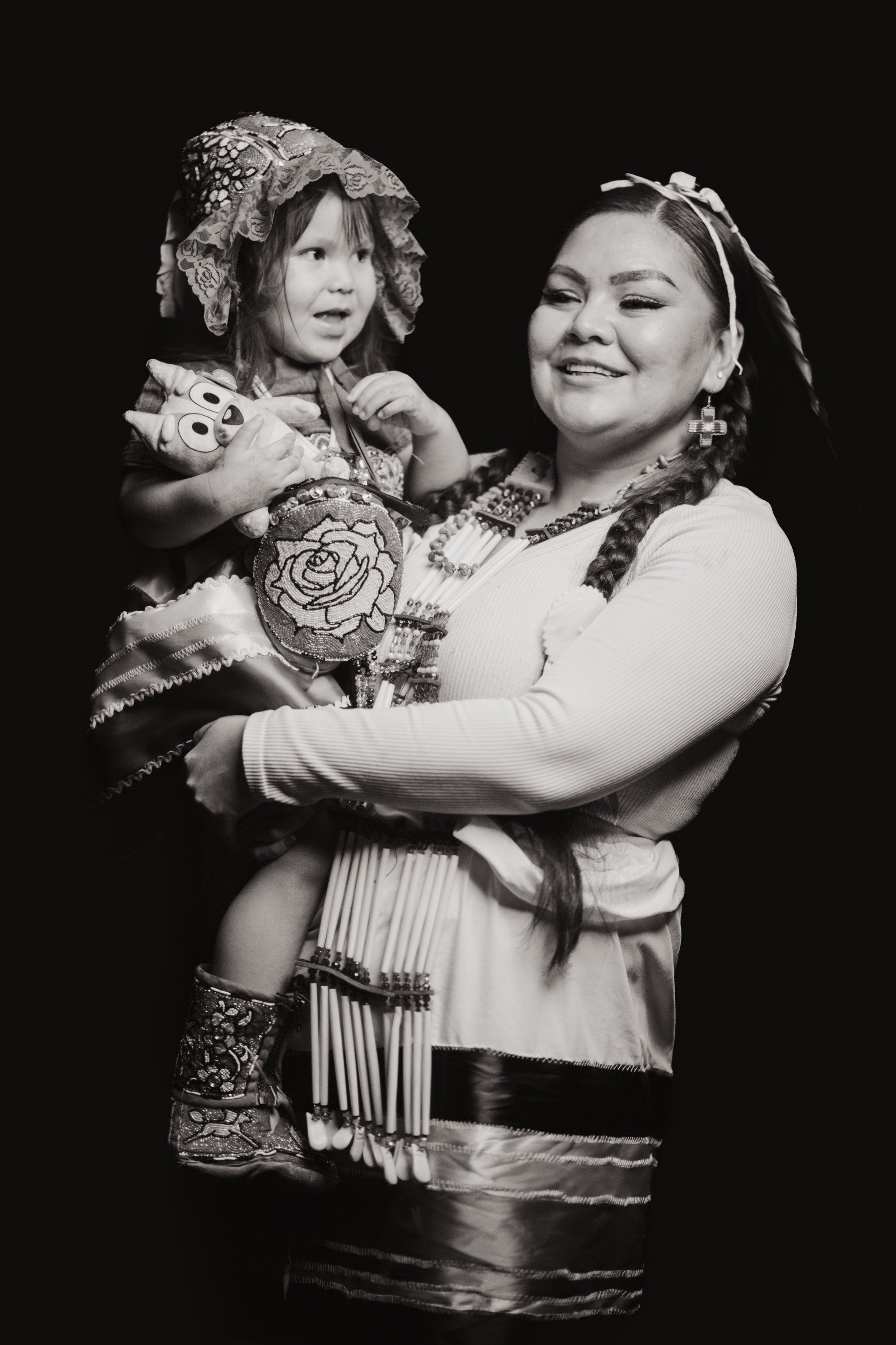 Woman and child in traditional Coast Salish regalia smile warmly against black background, child holding decorated rattle