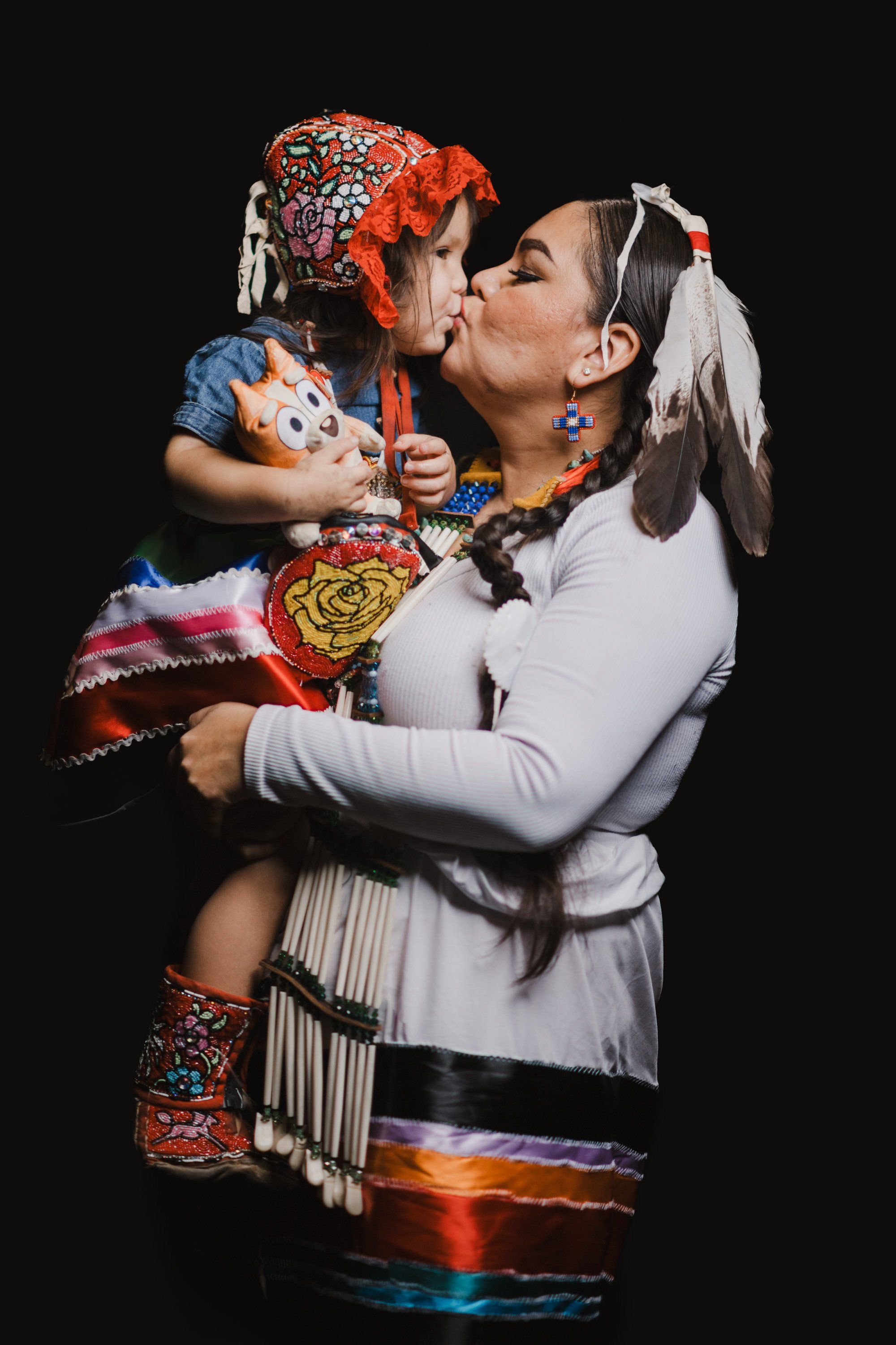 Woman in white regalia kisses child in colorful Coast Salish clothing against black background
