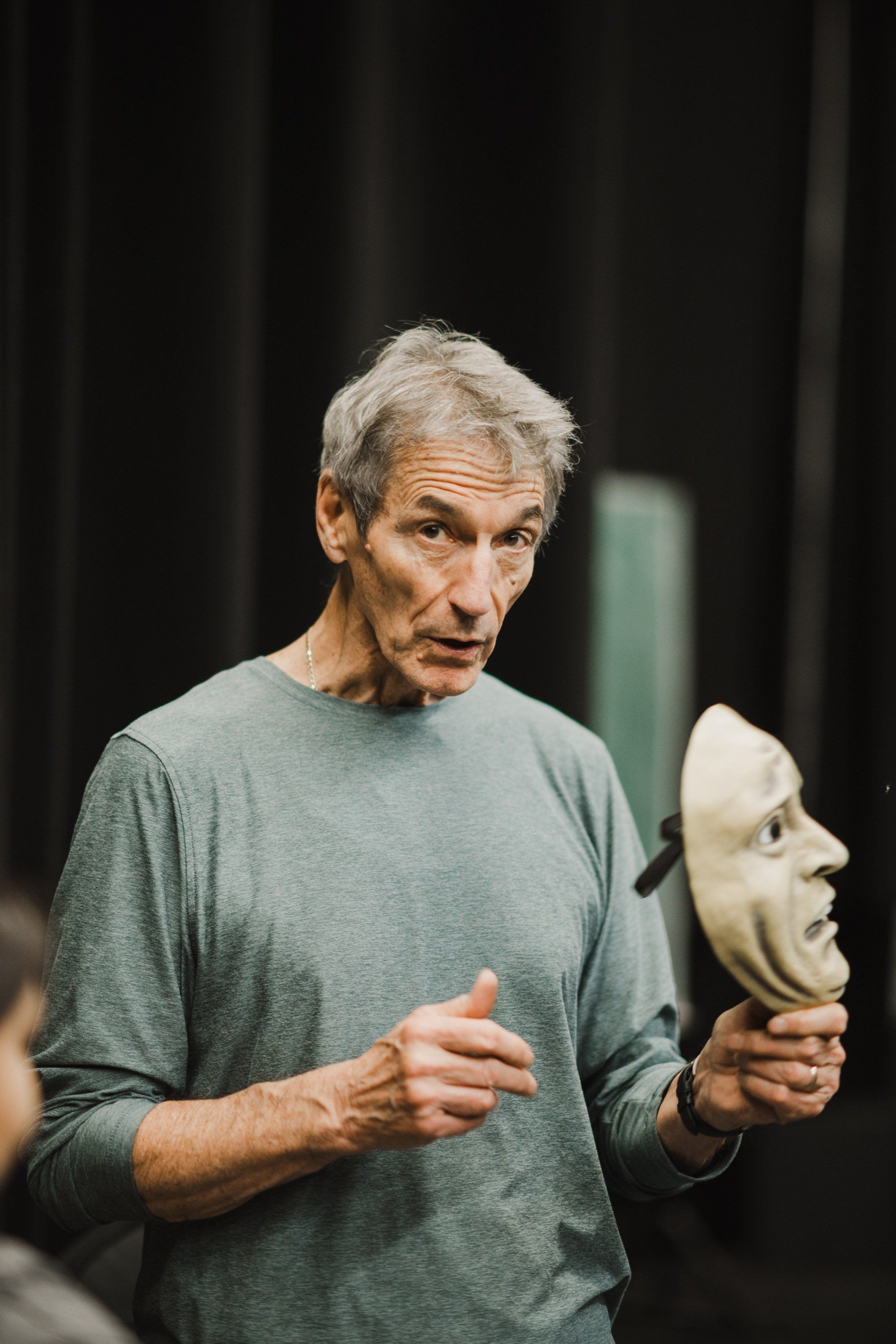 Elder holding carved wooden mask while speaking, dark curtain backdrop, candid moment of cultural storytelling