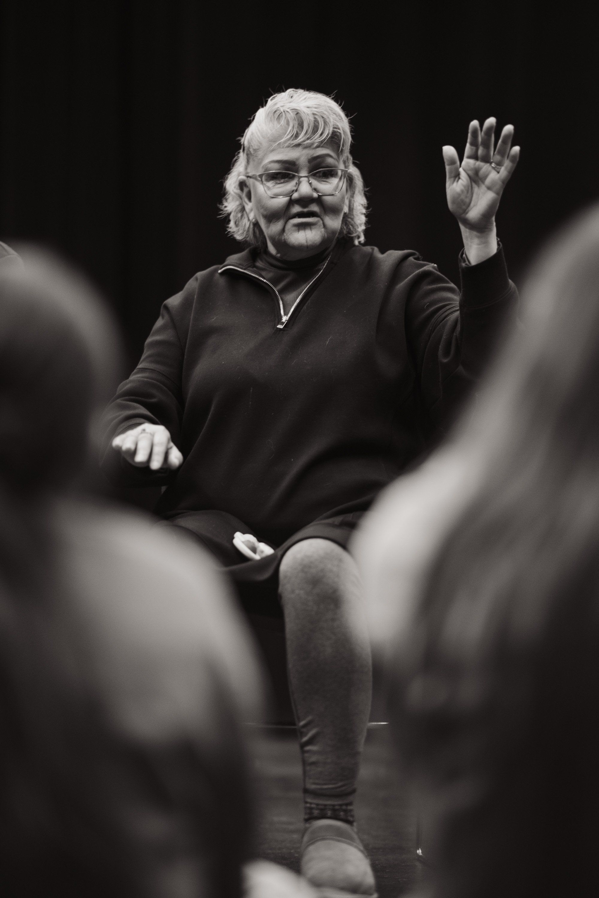 Elder in glasses and dark jacket raises hand while speaking to audience members in foreground, black and white portrait