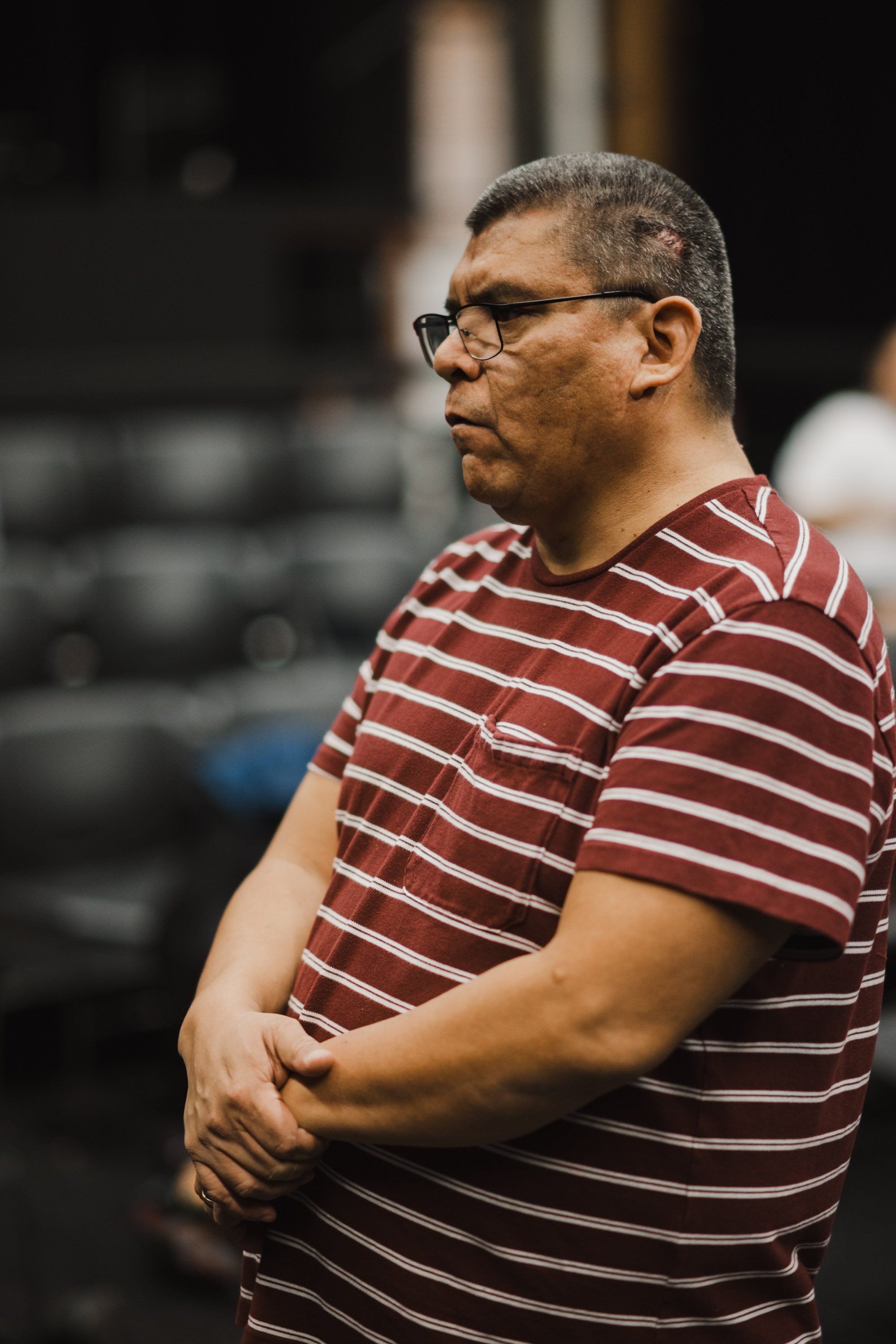 Middle-aged man in striped burgundy and white shirt and glasses, captured in profile against an indoor venue backdrop during what appears to be a cultural event
